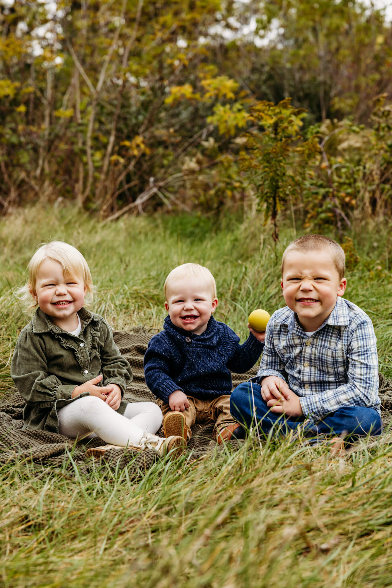 3 young siblings sitting together in the grass and smiling in a park in Door County.