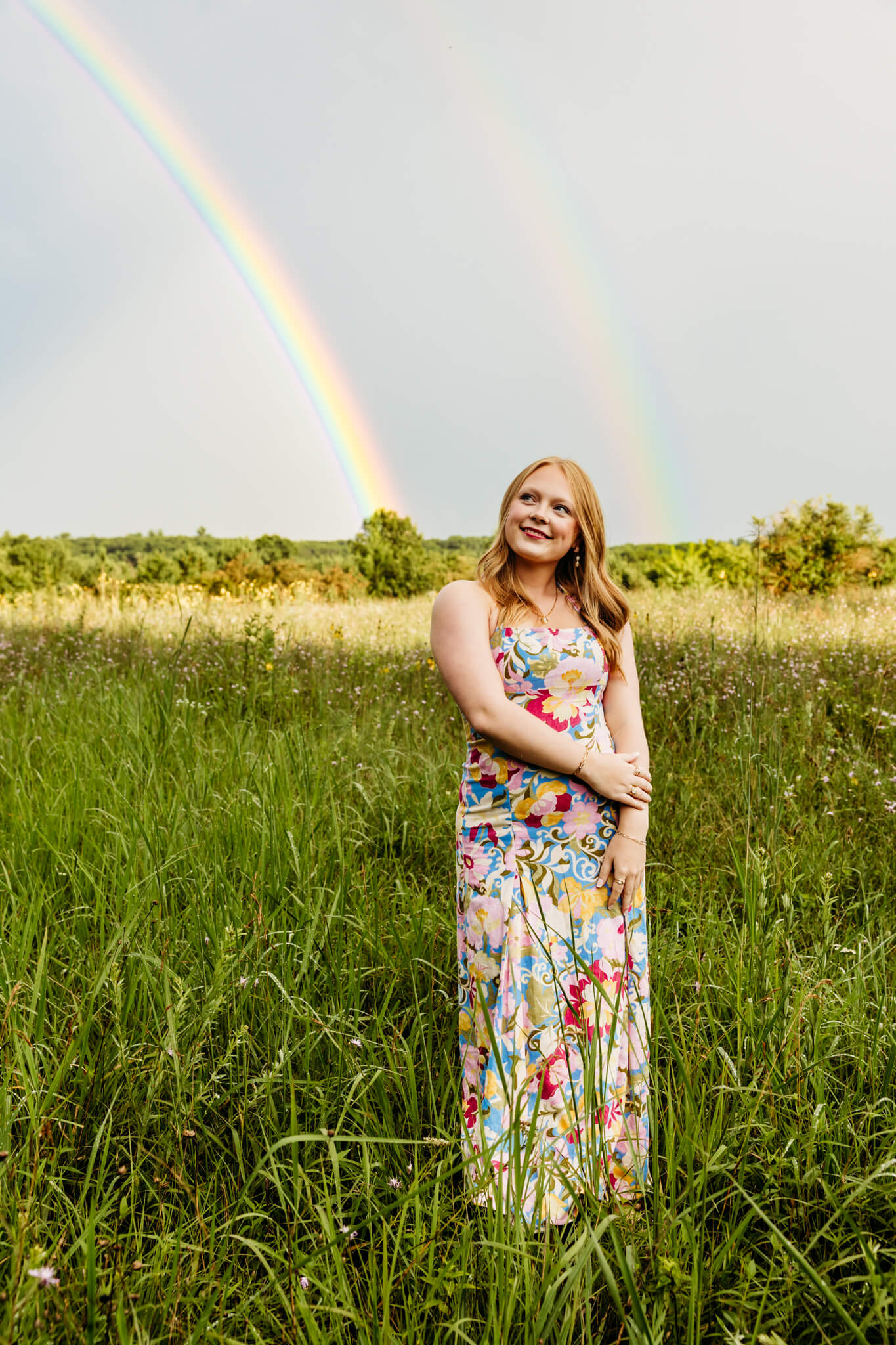 high school girl in a colorful dress standing in field as two rainbows appear behind her near Door County.