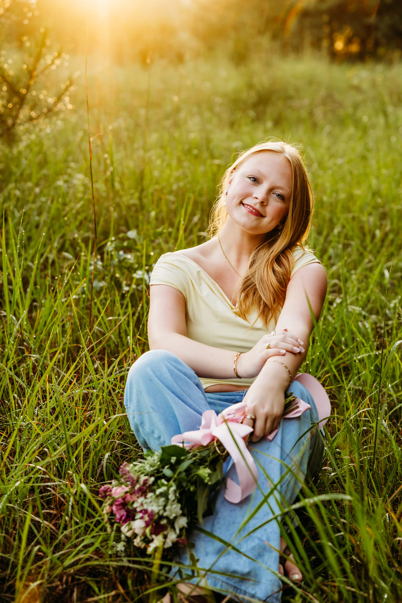 Young lady in a yellow top and jeans holding a bouquet while sitting in tall grass. 