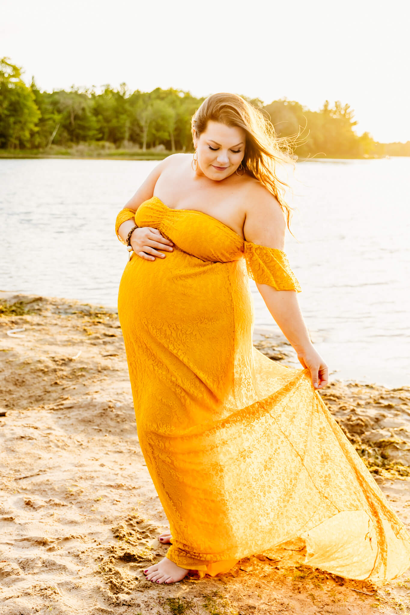 mama to be playing with her yellow lace dress as she walks along the beach with the sun setting behind her