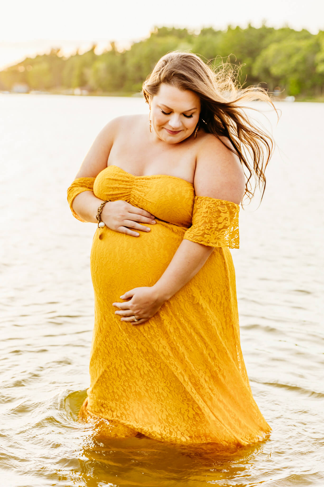 stunning expecting mother holding her baby bump as she looks down at her belly and the wind blows in her hair while standing in a lake near Green Bay
