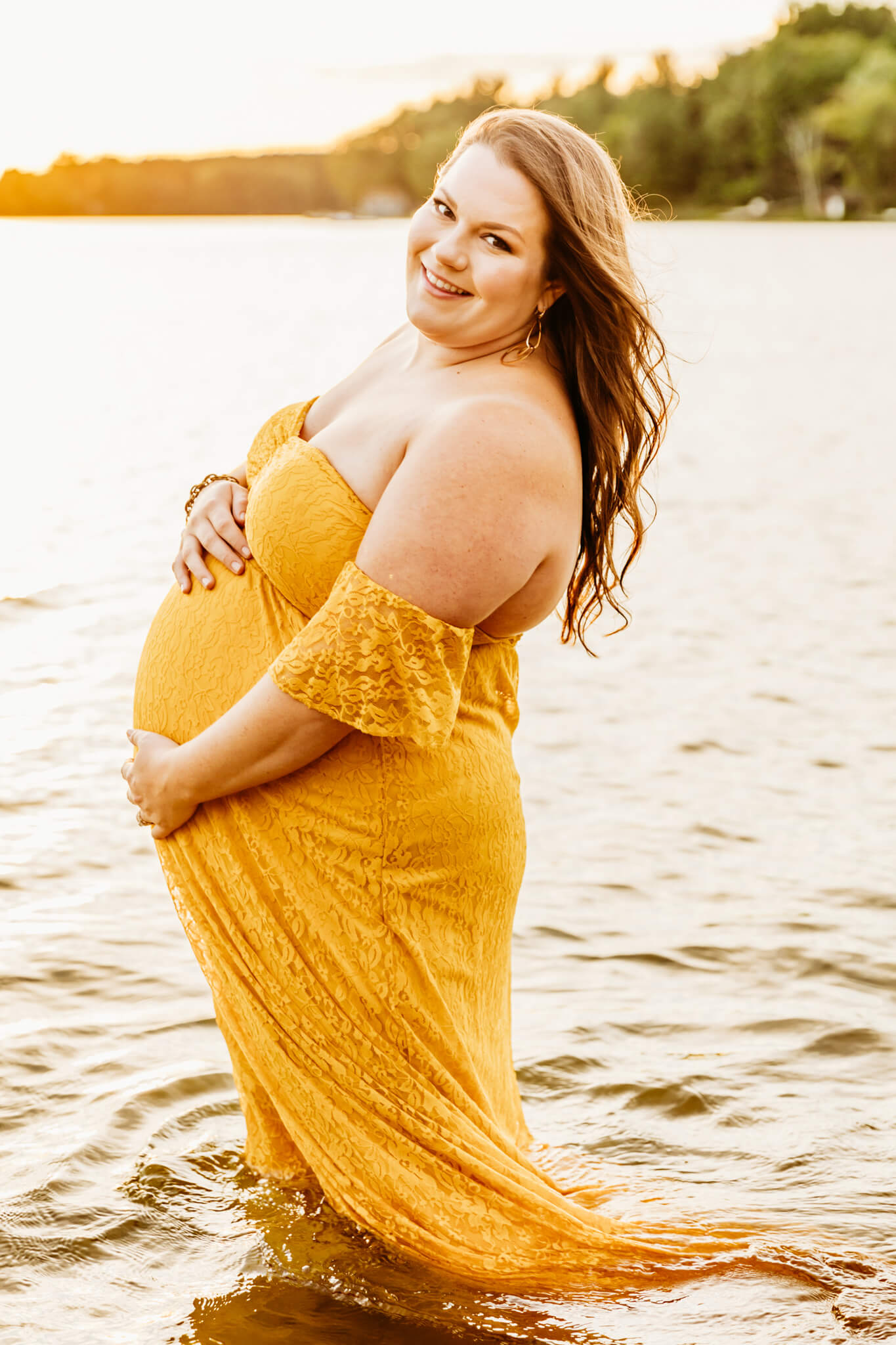 pregnant woman in a yellow lace dress standing in a lake and holding her baby bump at sunset 
