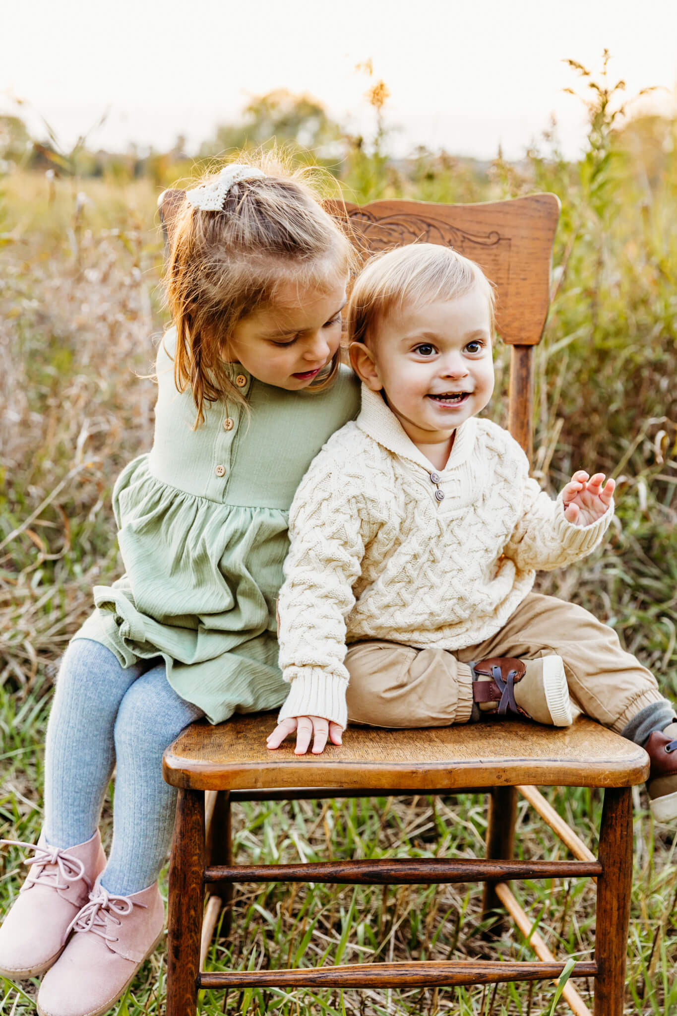 Little girl in a green dress looking at her younger brother in a cream sweater while sitting on a wooden chair together