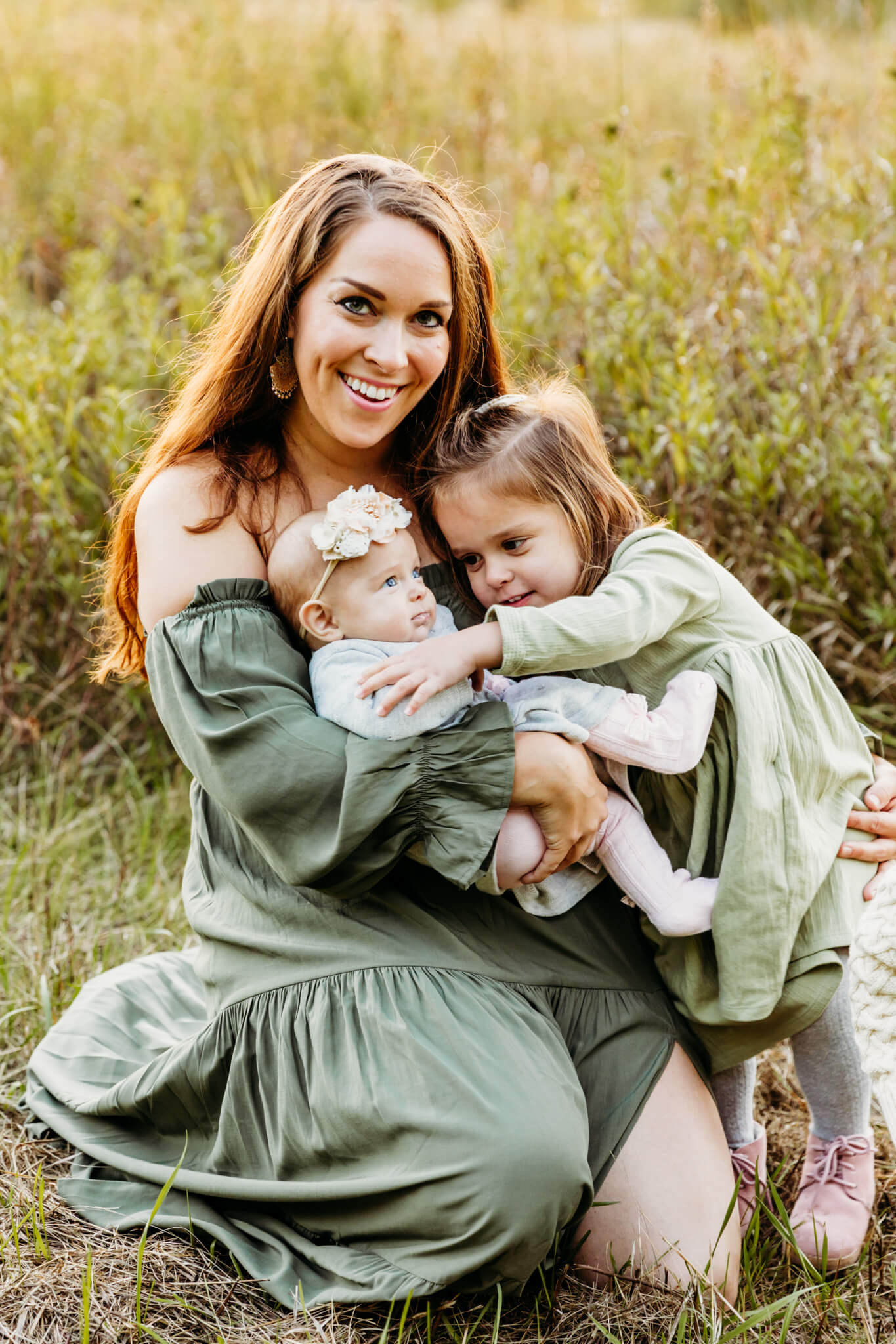 beautiful mother in a sage green dress smiling as she holds her little daughters in a field near Oshkosh