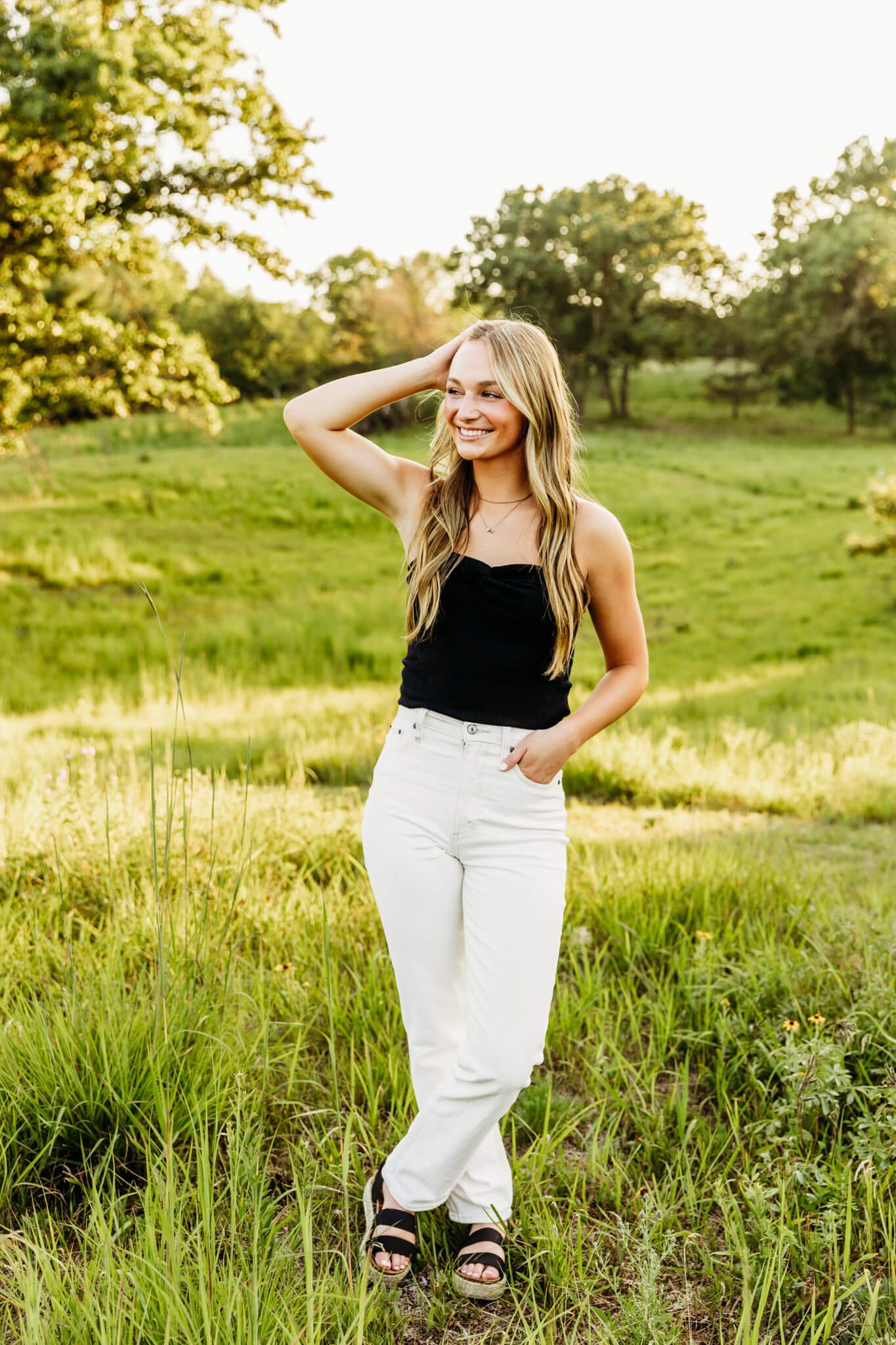 beautiful high school girl in white pants and a black top standing with her ankles crossed, one hand in her pocket and one in her hair as she looks out into the distance smiling
