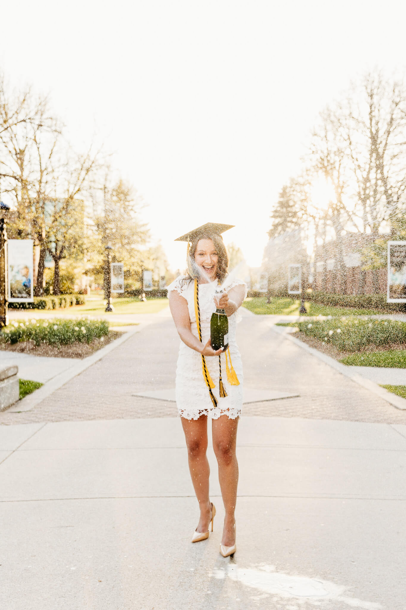 college grad in a white lace dress wearing her graduation cap while popping champagne and spraying it for a blog post about colleges in Green Bay.