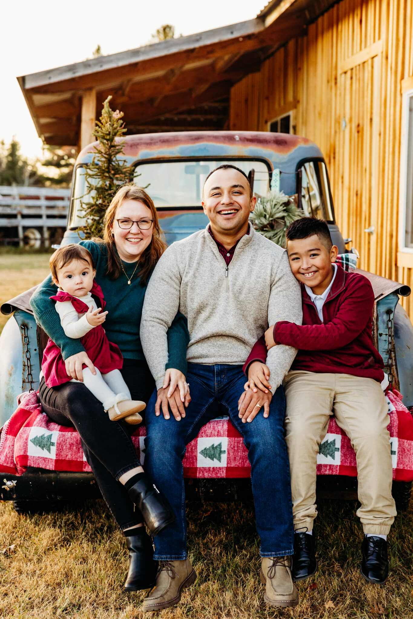 Mom and dad sitting on a vintage truck bed with their two children and smiling for their family photos at a Christmas Tree Farm.