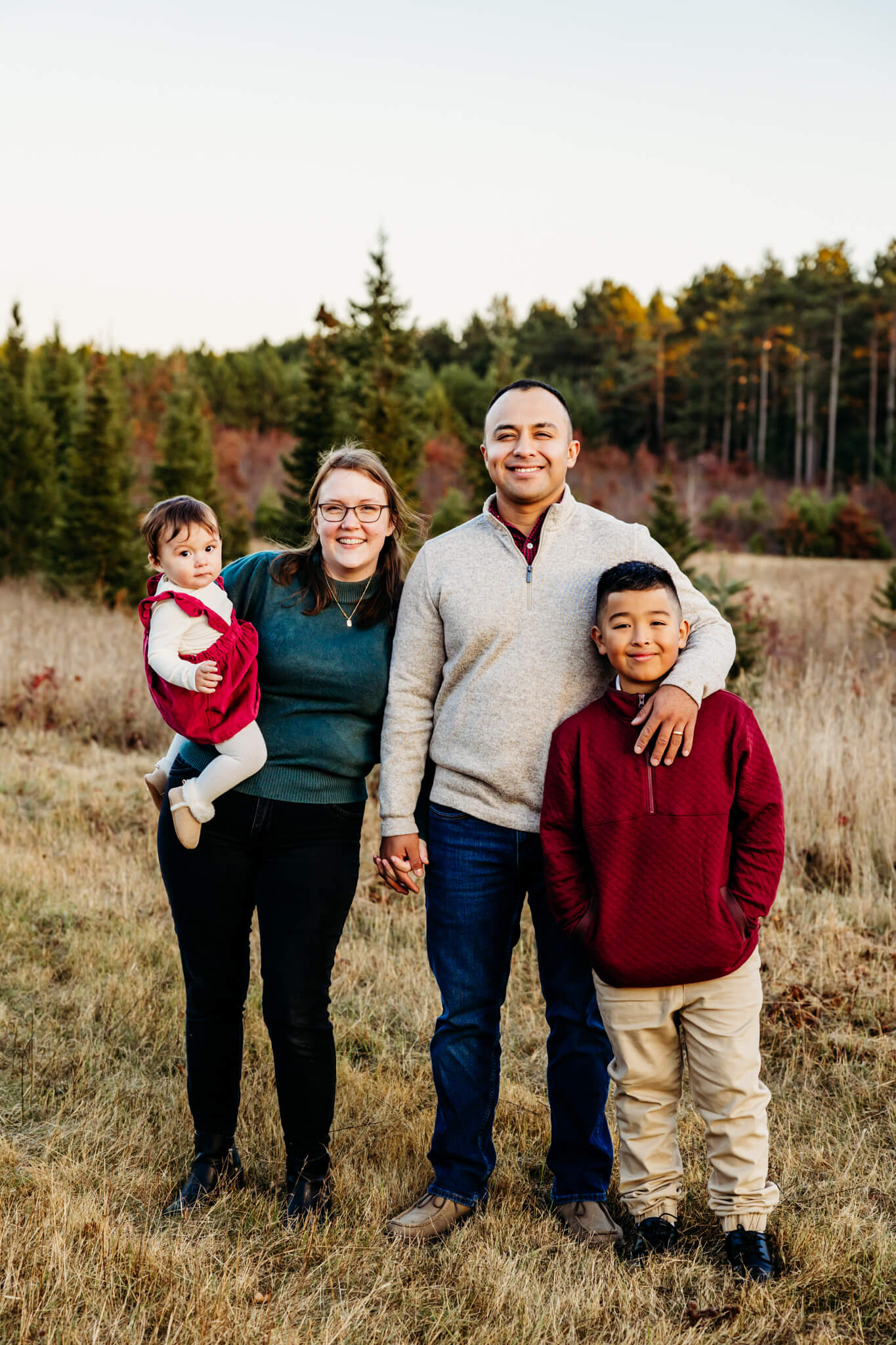 mother and father snuggling with their two children as they stand in front of Christmas trees at Balsam Creek Lane Tree Farm in Black Creek, WI