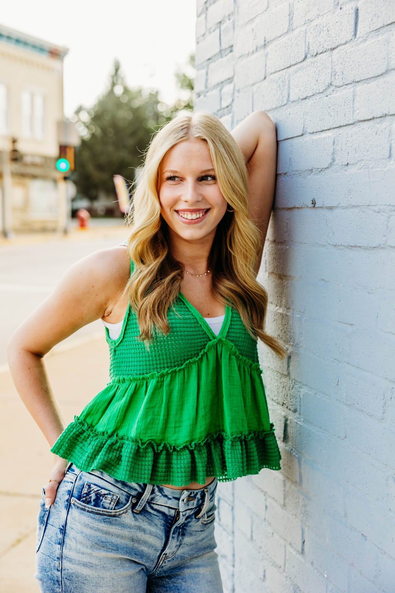 Blonde teen girl in a kelly green top and jeans leaning against a blue brick wall.
