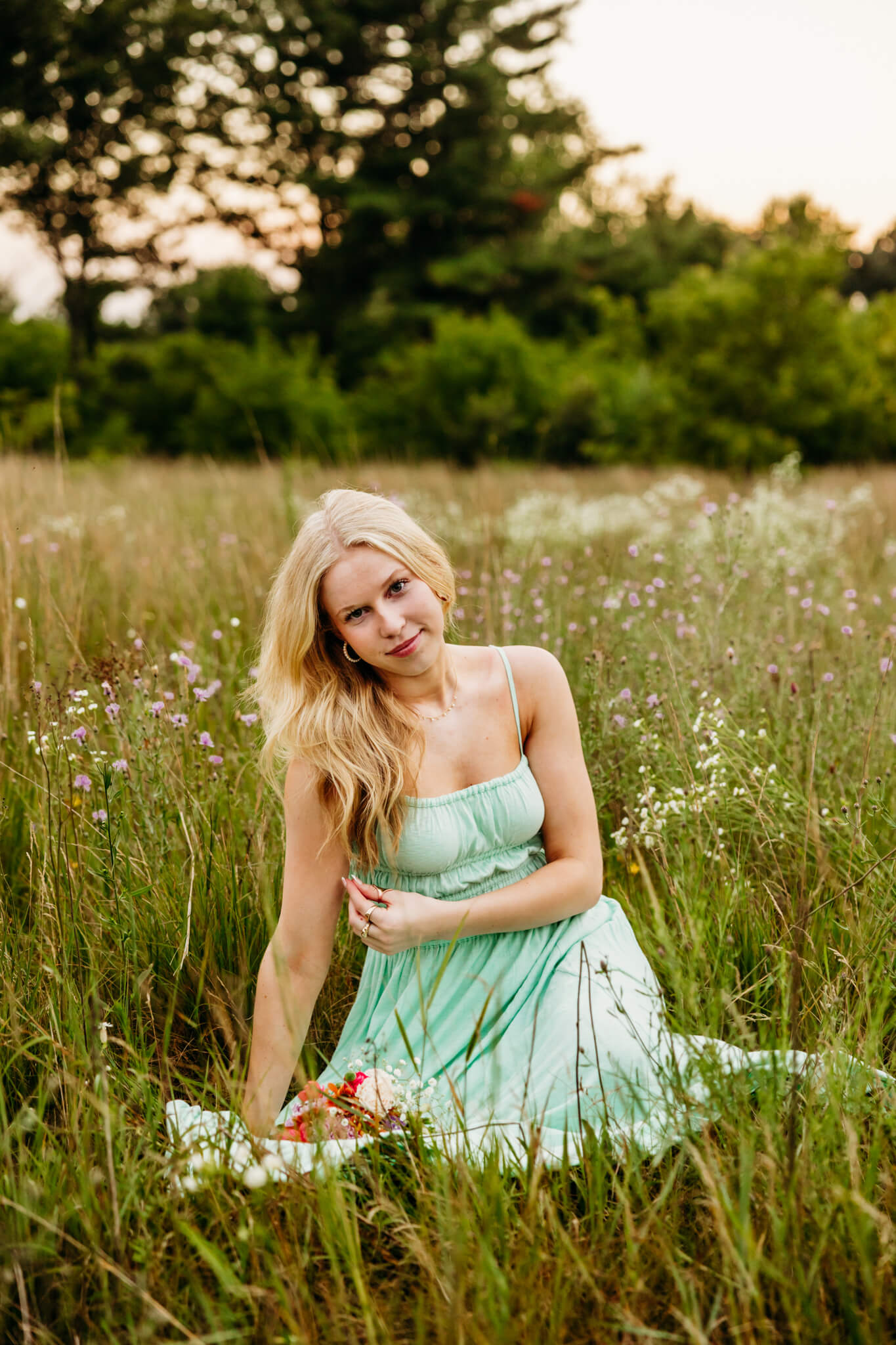 High school girl in a green dress sitting in a flower field and playing with her hair for her senior pictures.