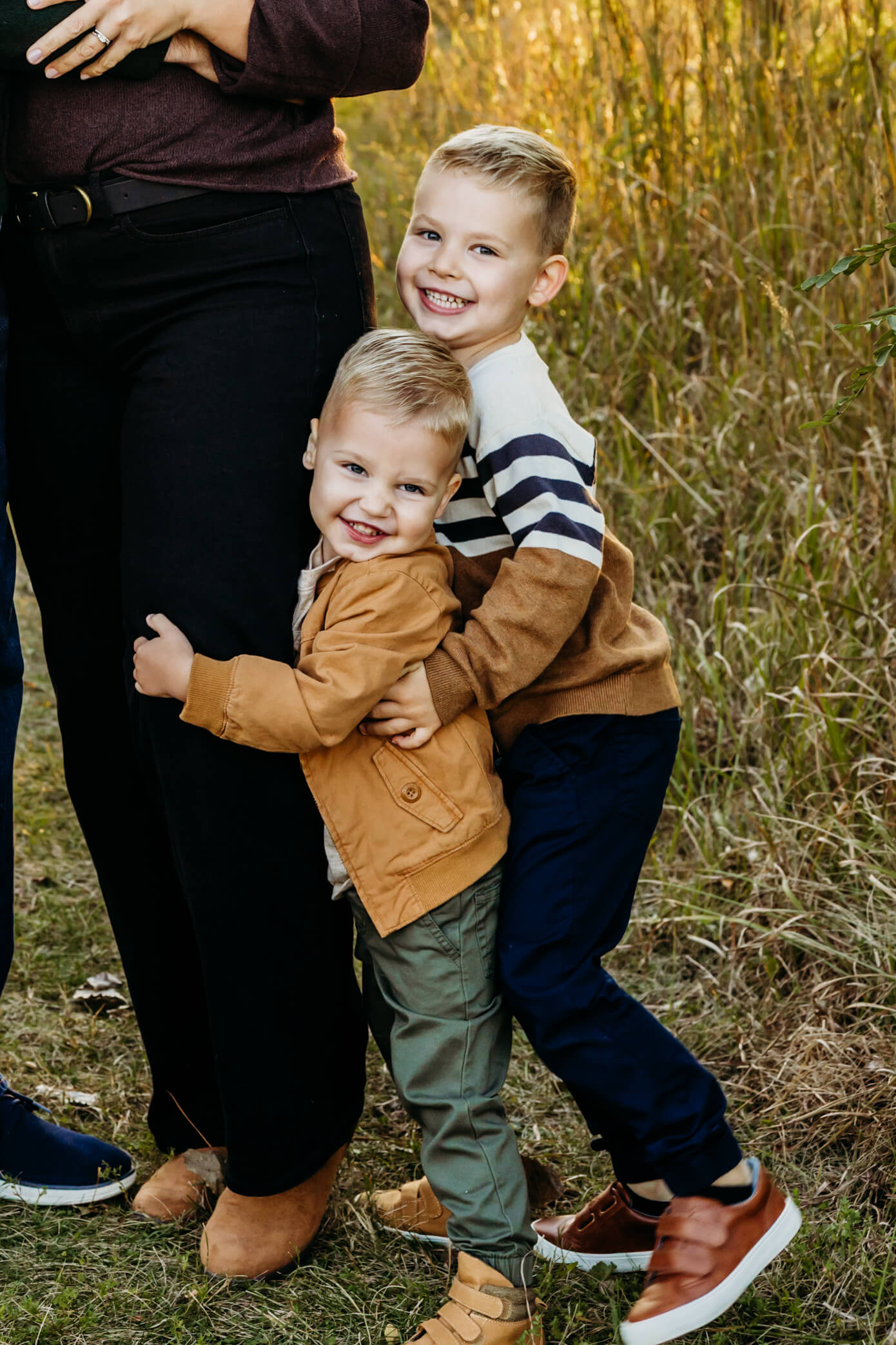 Two young brothers hugging each other and making silly faces as they hug their mom's leg.