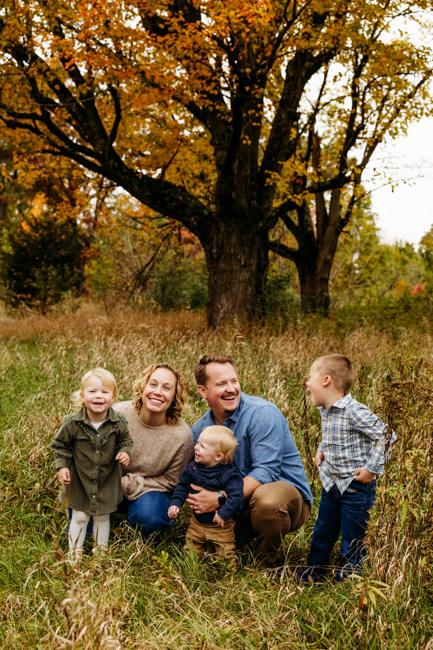 mother and father kneeling with their 3 younger children as they laugh and play in a field near Oshkosh WI