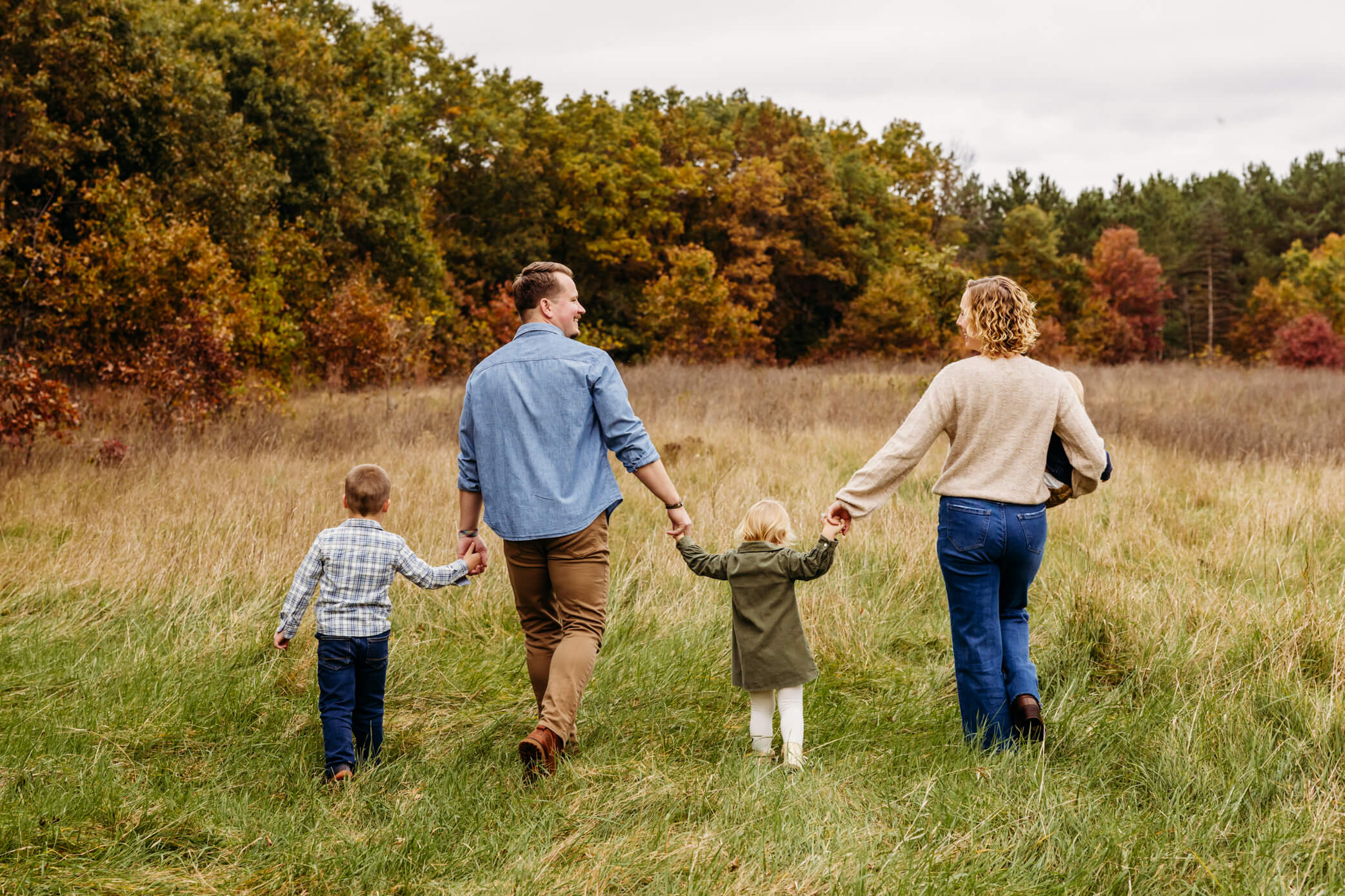 mom and dad holding hands with their young children as they walk in a fall colored field together looking at each other.