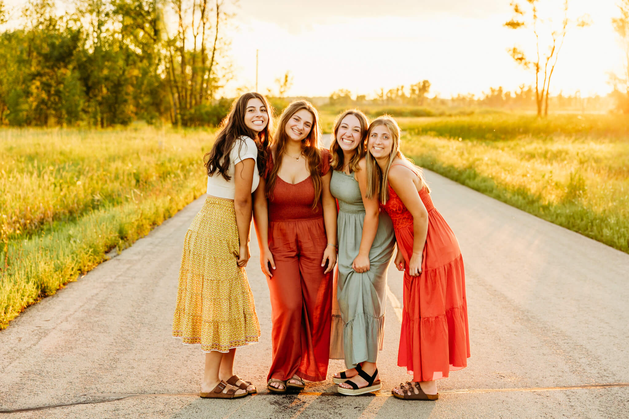 four high school senior girls standing together while dressed in colorful boho outfits on a backroad at sunset near Oshkosh