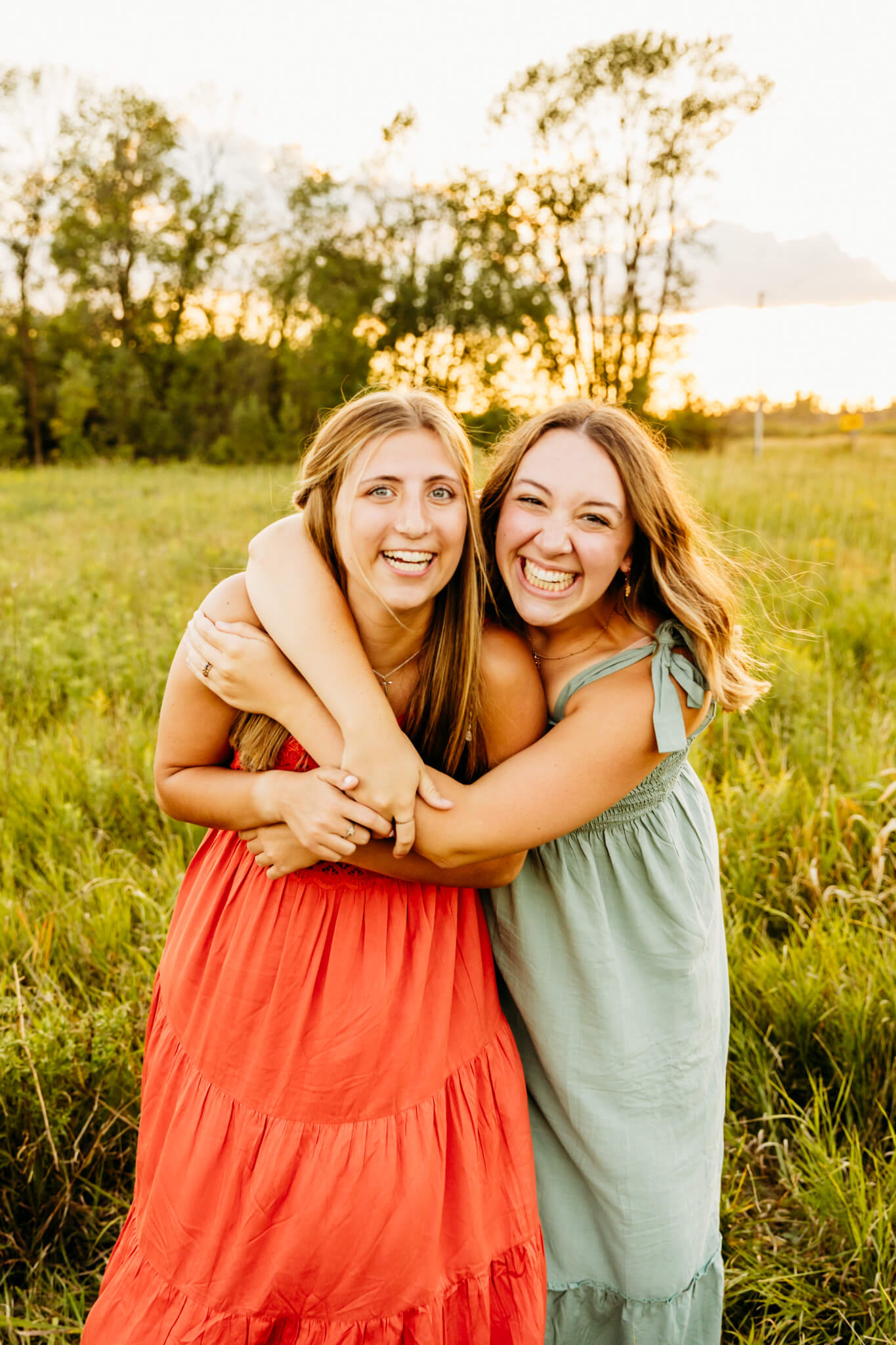 teen girl in a green dress hugging another teen girl in an orange dress in a field at sunset as they both laugh