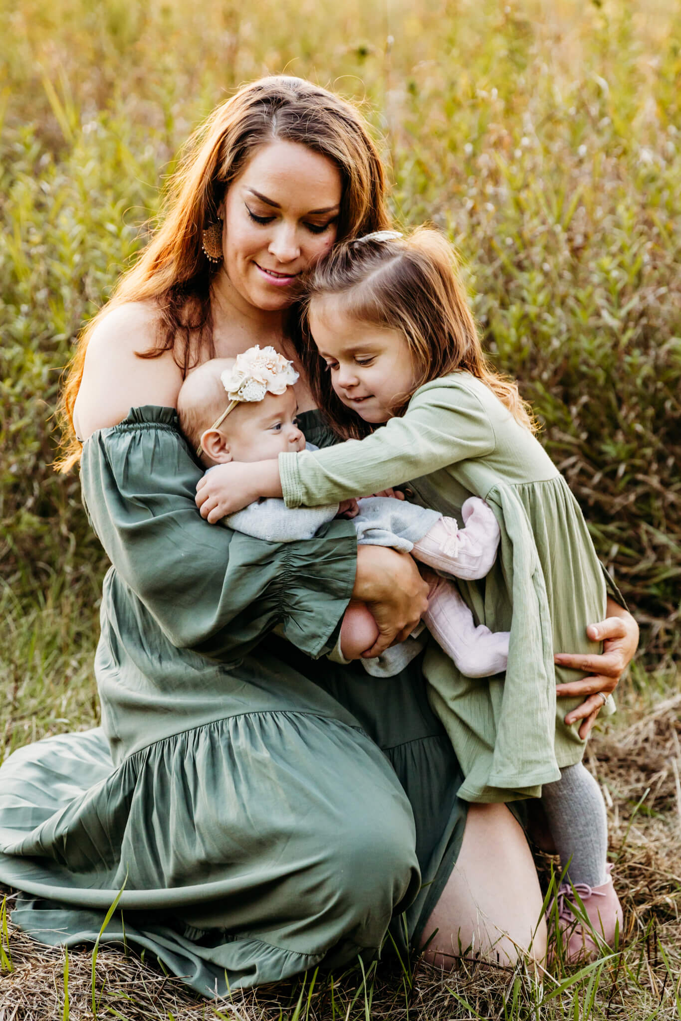 mama in a green dress kneeling down and hugging her young daughter and holding her baby girl