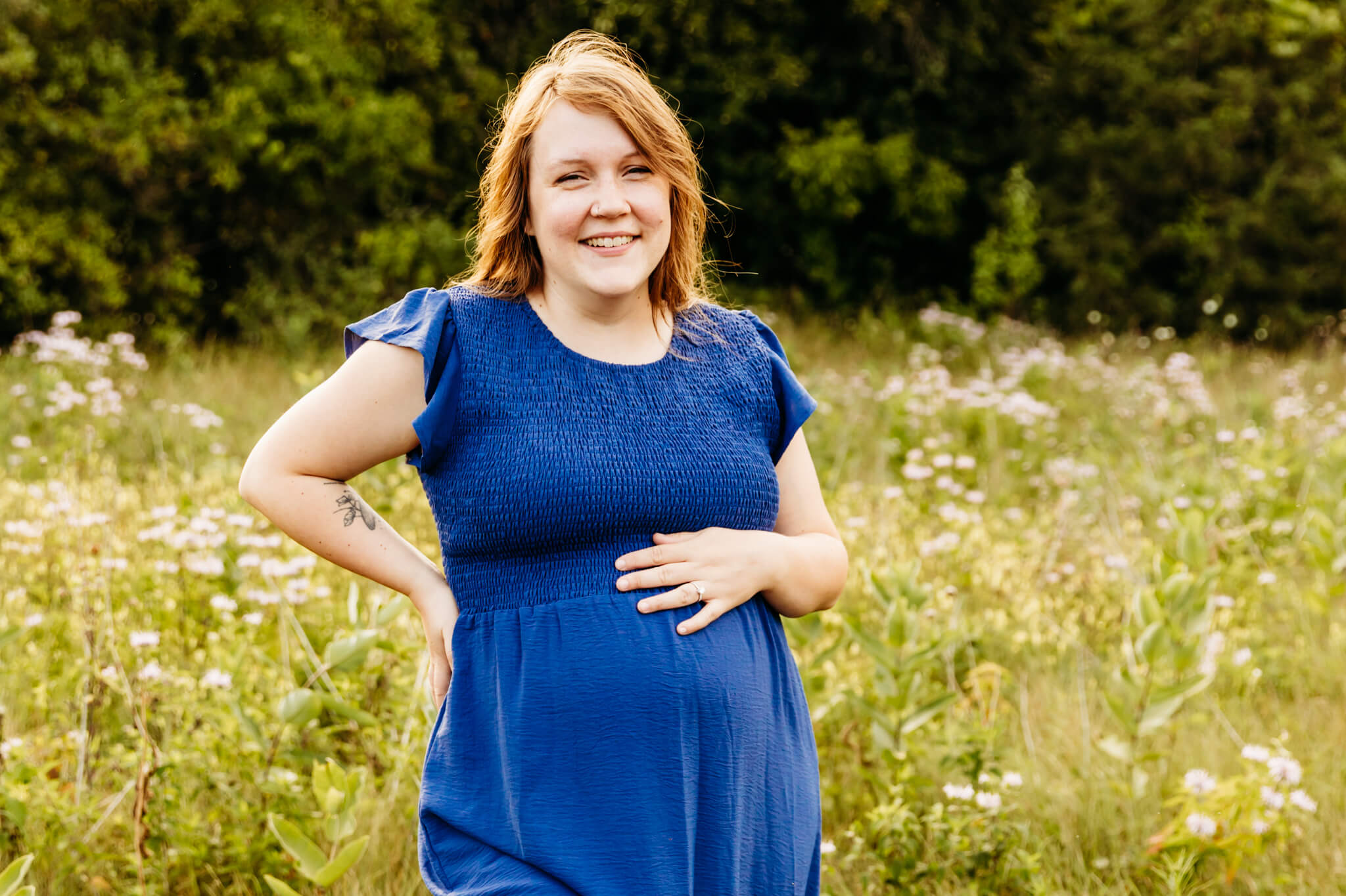 gorgeous mama to be with red hair in a blue dress standing with one hand on her hip and the other on her baby bump in a flower field for a blog post about spas in Oshkosh