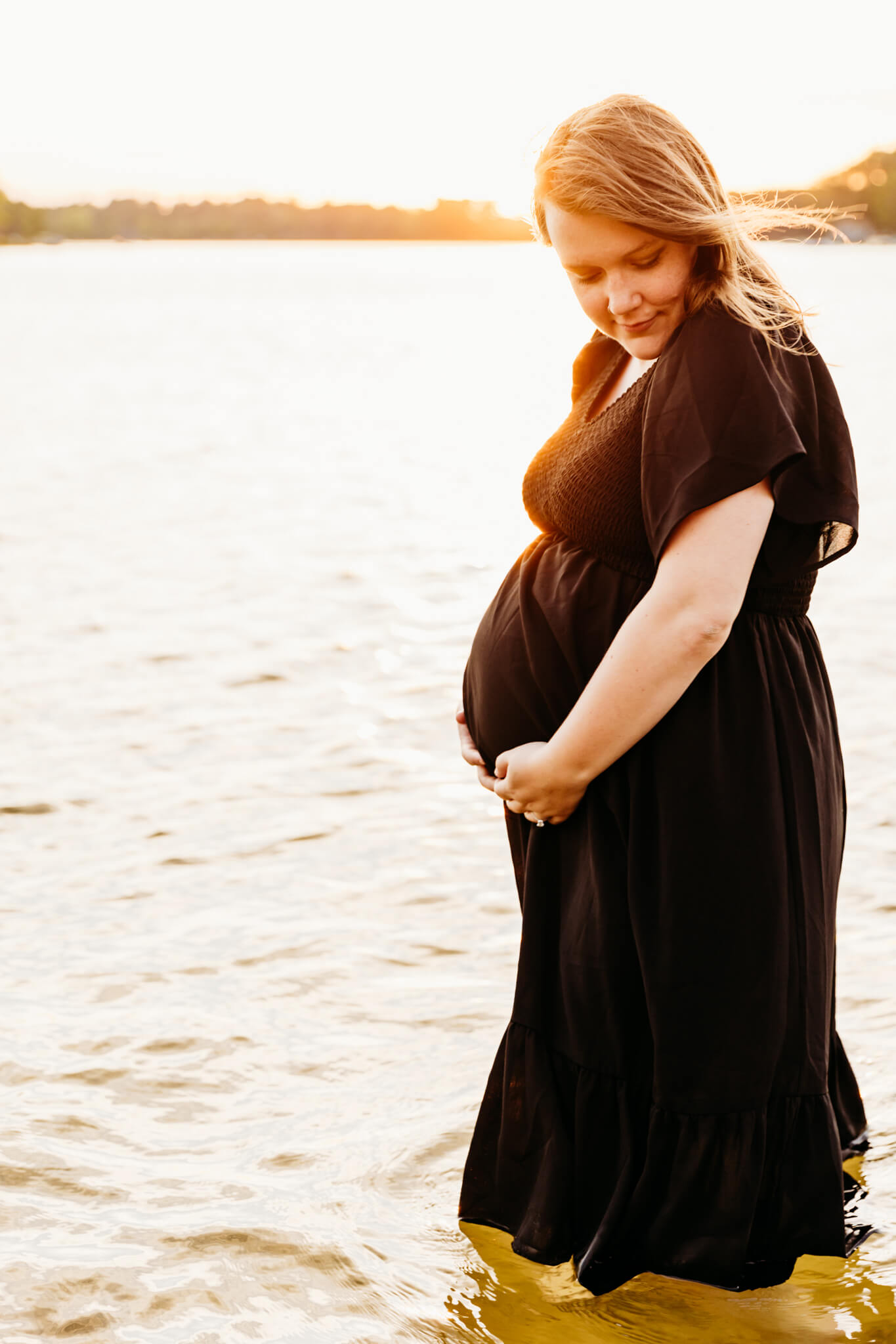beautiful pregnant woman with red hair standing in a lake in a black dress and holding her baby bump as she looks down at her arm
