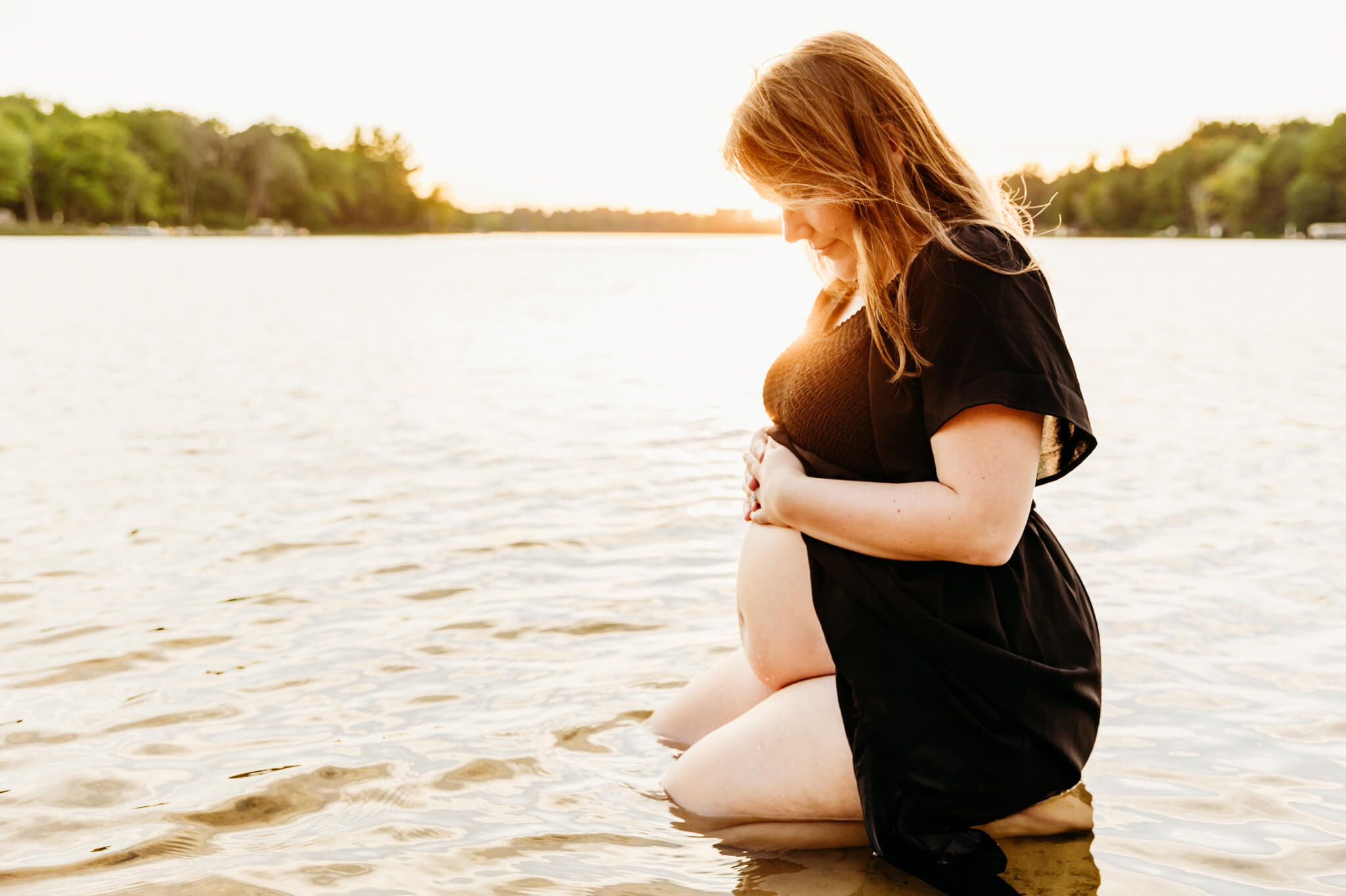 expecting mama in a black dress kneeling in a lake and looking down at her baby bump as the sun sets behind her