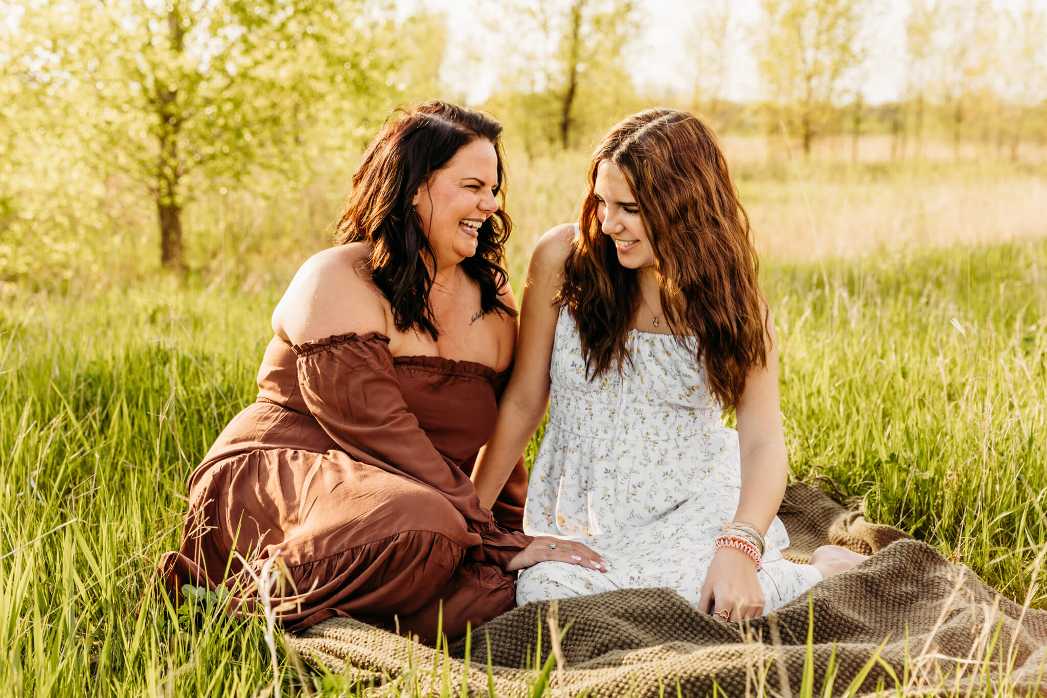 mom in a brown dress and teen daughter in a white dress laughing as they snuggle together on a blanket 