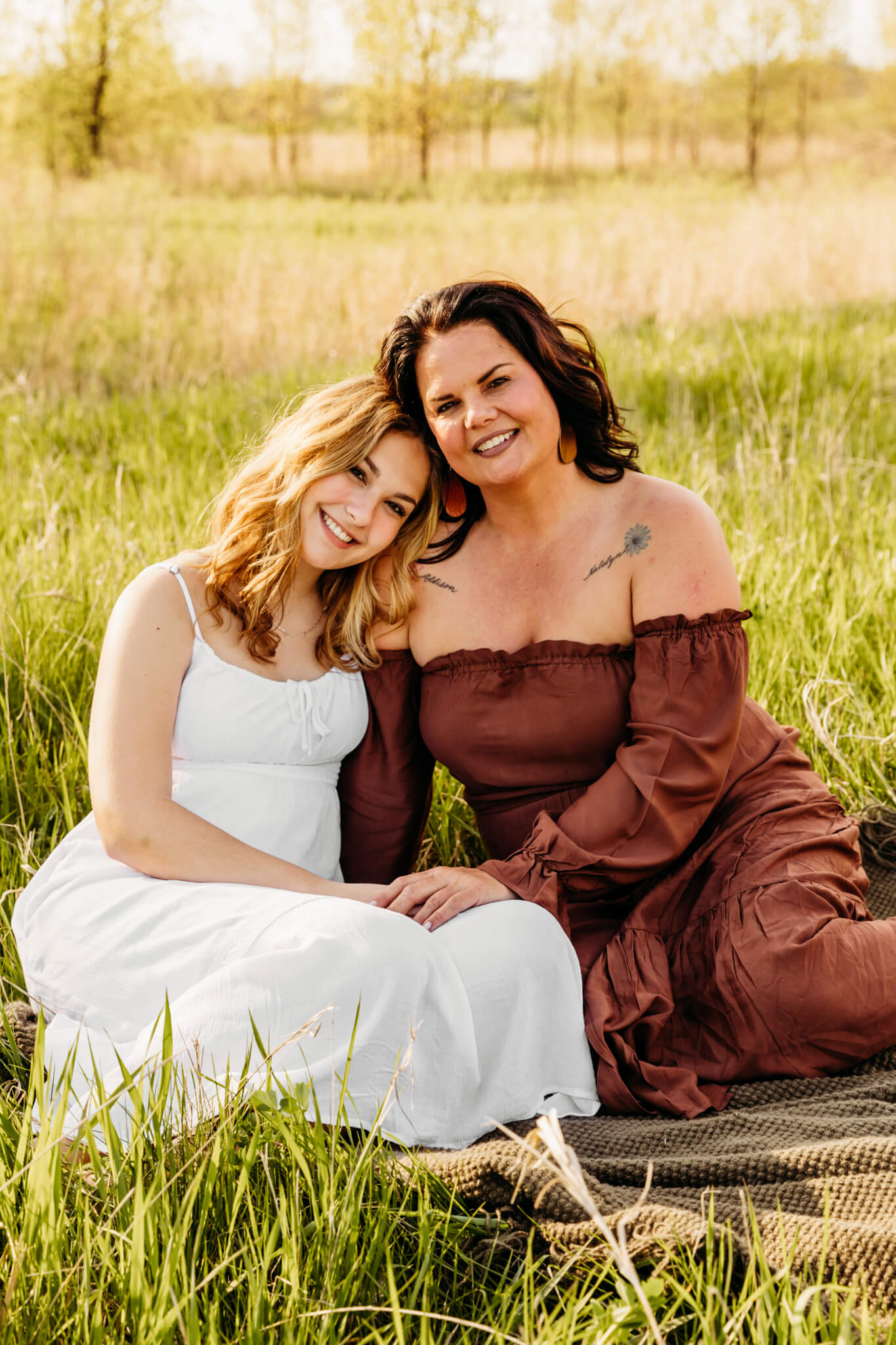 teen daughter resting her head on her moms shoulder as they sit in a grassy field