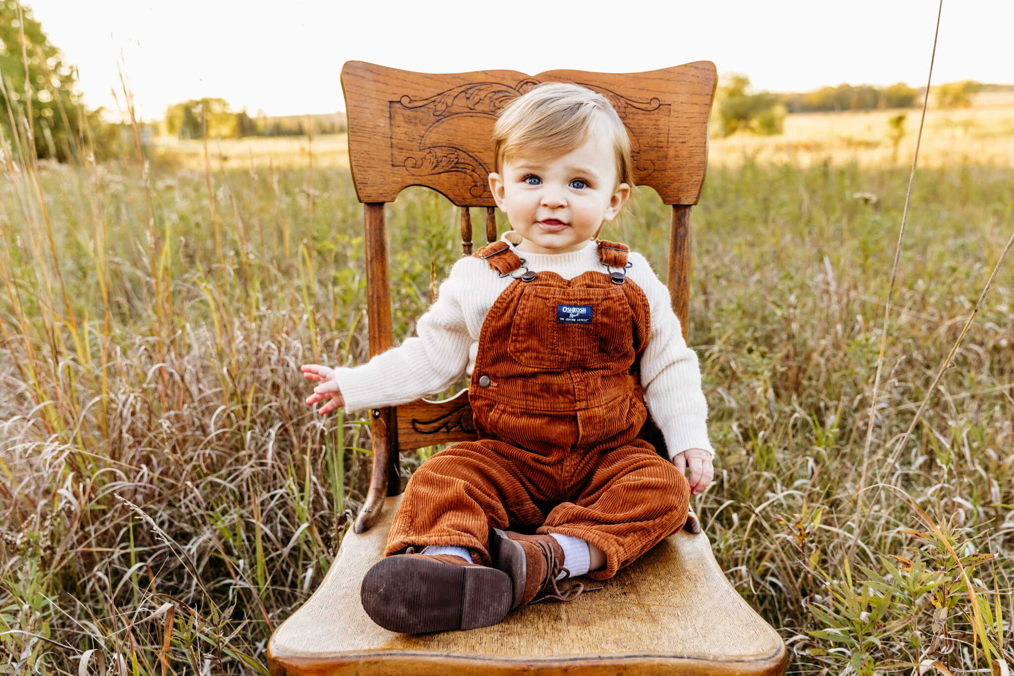 baby boy in orange overalls sitting on a brown wooden chair in a field for a blog post post about swim lessons in Appleton WI