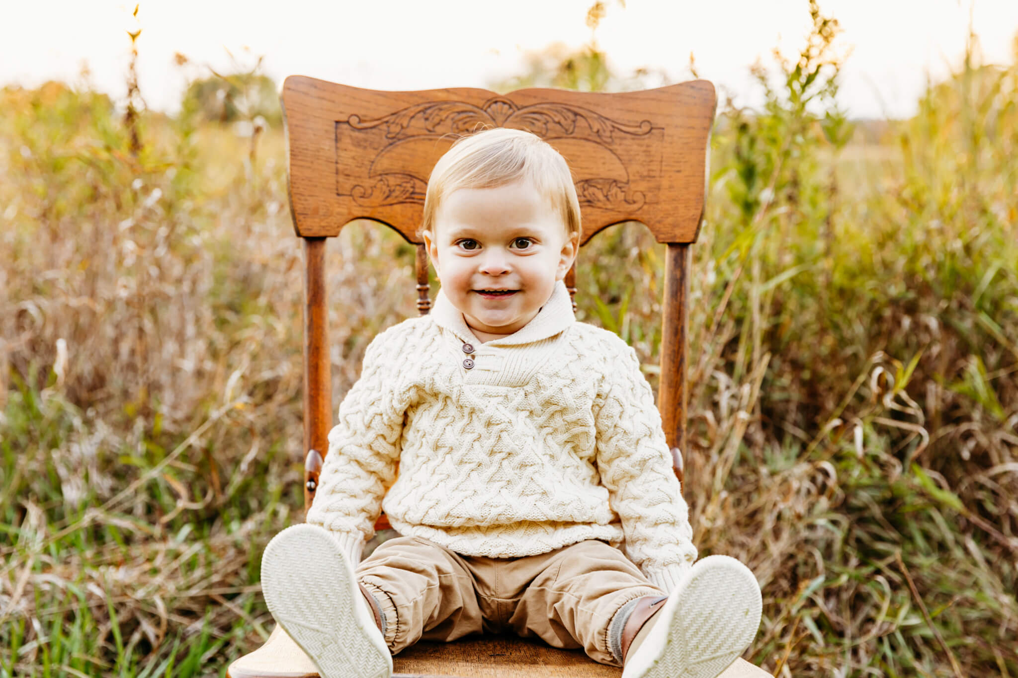 baby boy in a cream color sweater smiling and sitting on a chair in a grassy field near Green Bay