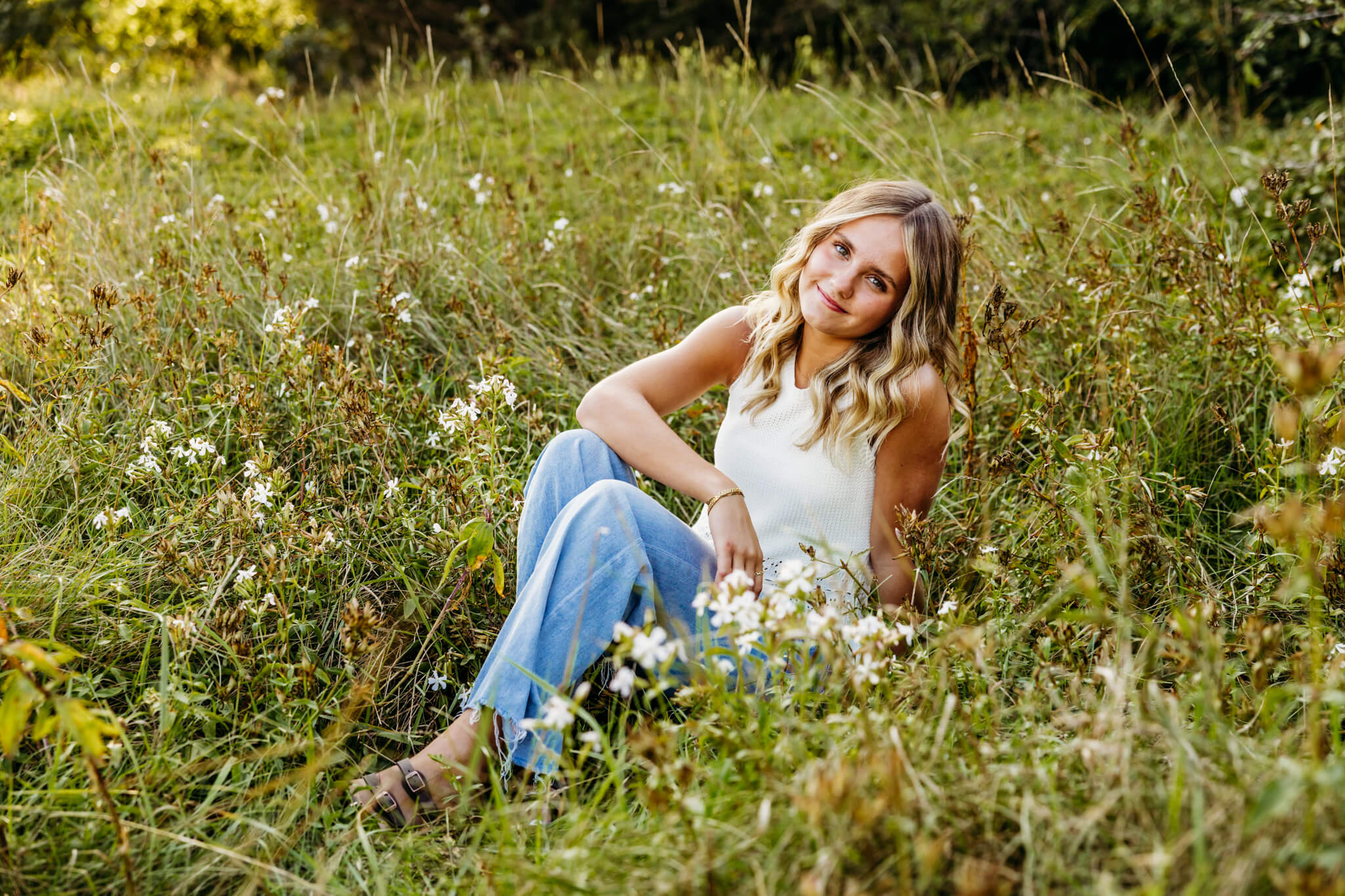 Stunning teen girl sitting in a grassy field in a white tank top and jeans in a park near Fond du Lac.
