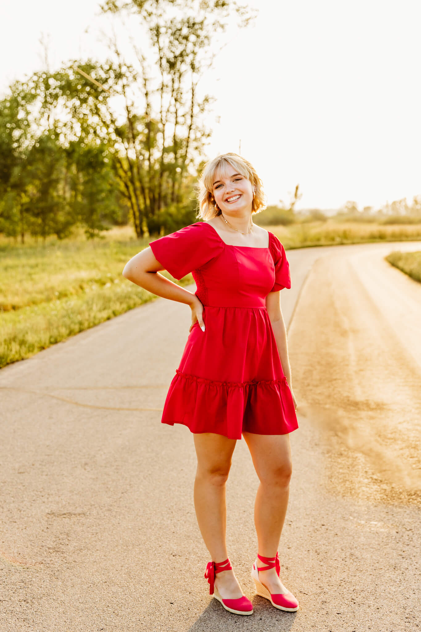 beautiful teen girl in a red dress and red wedges standing on a back road at sunset and smiling