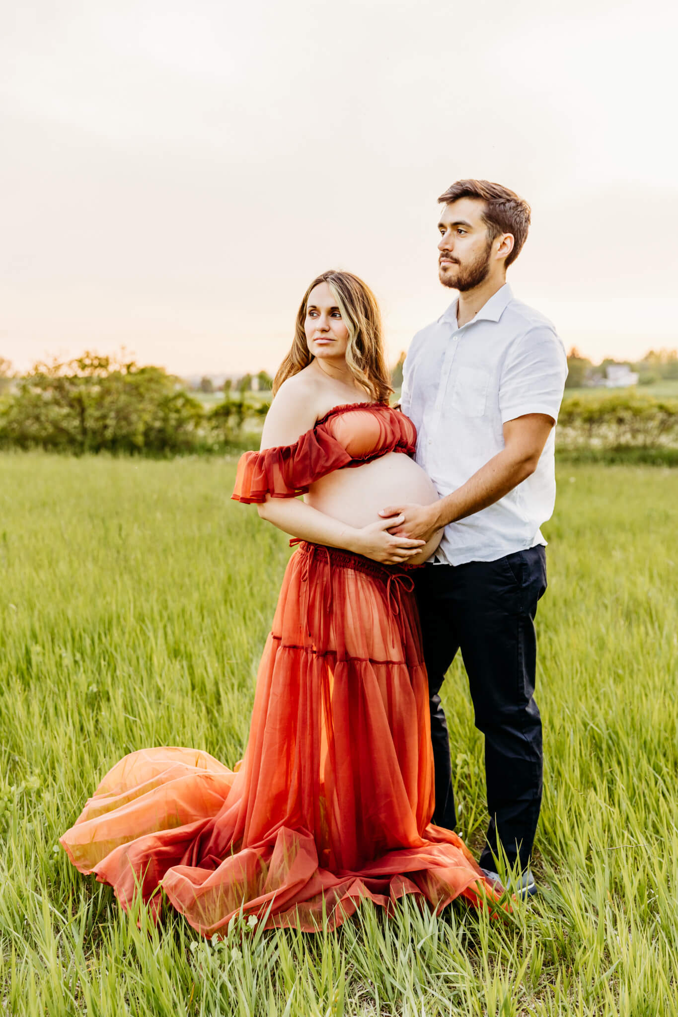 husband and wife holding each other and embracing their baby bump as they look off into the distance