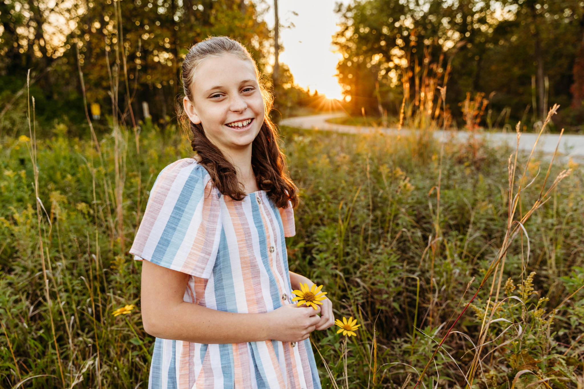A young girl in a stripe dress picks yellow wildflowers in tall grass of a park field at sunset