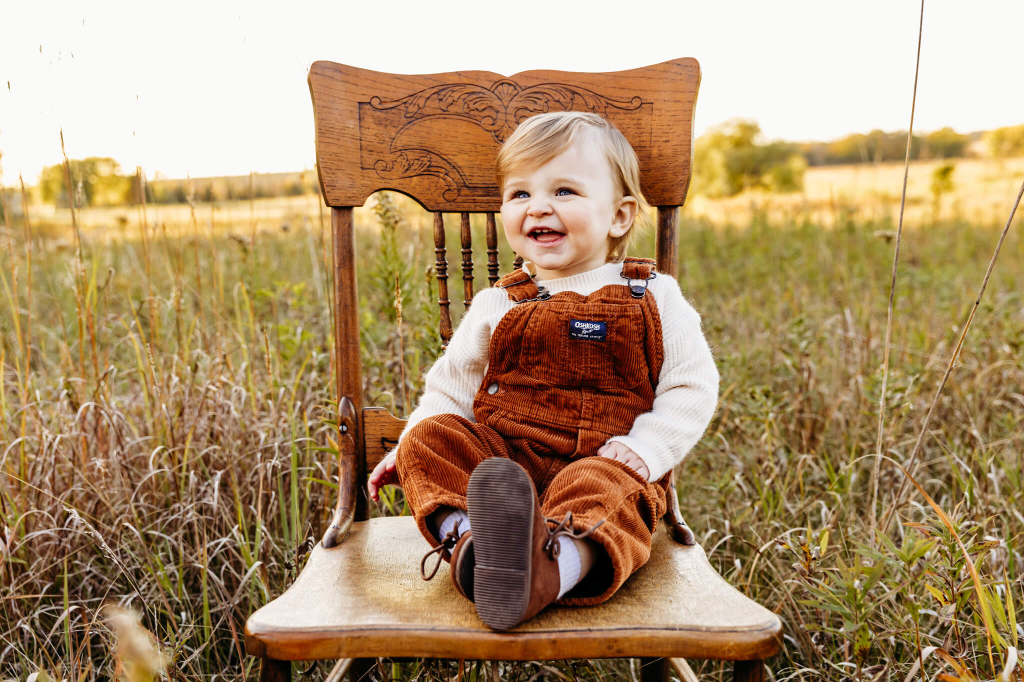 toddler in overalls sitting on a wooden chair in a field at sunset in neenah wi