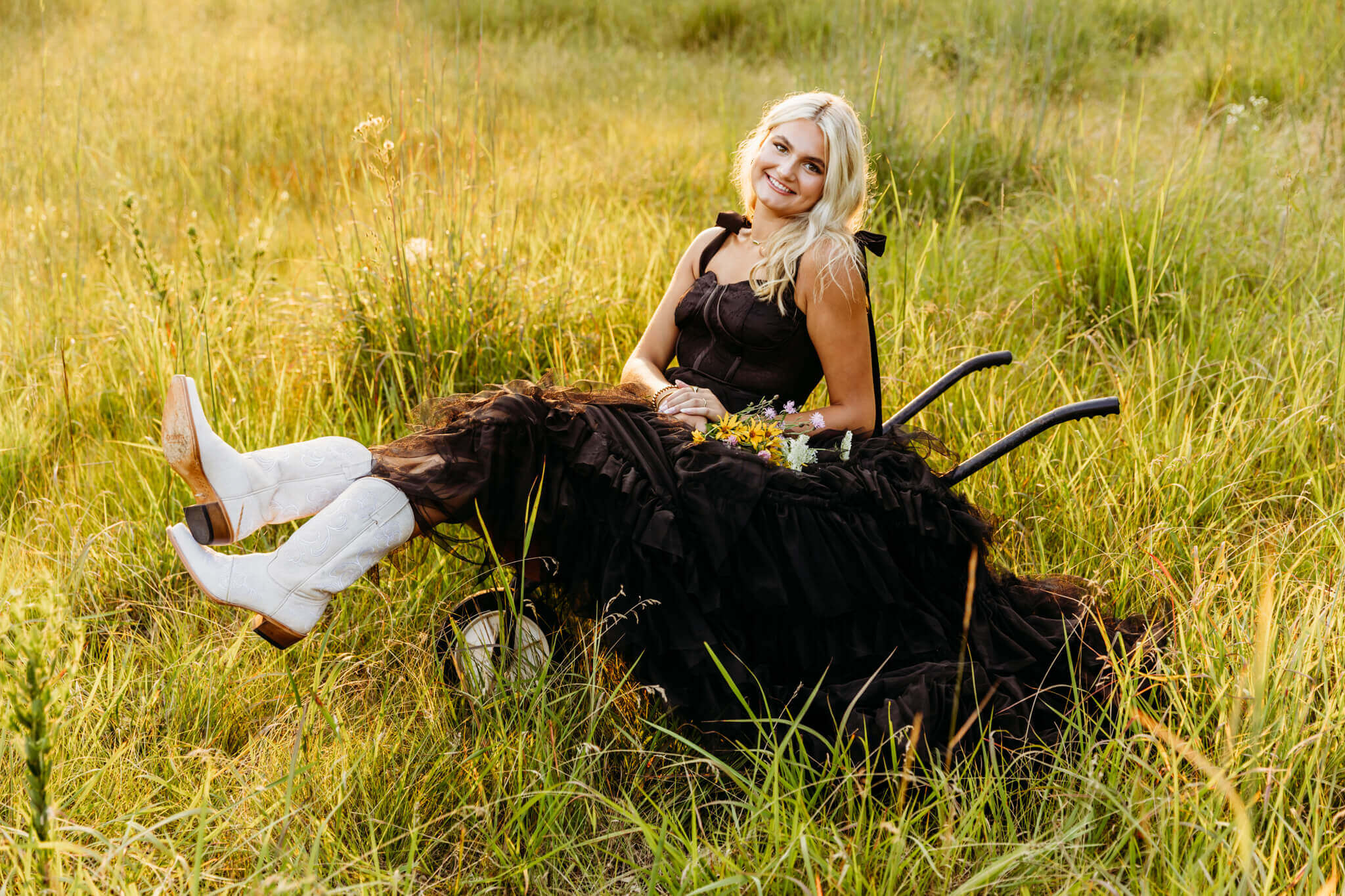 high school senior girl in a black gown, wearing cowboy boots sitting in a wheelbarrow in a field near Oshkosh, WI