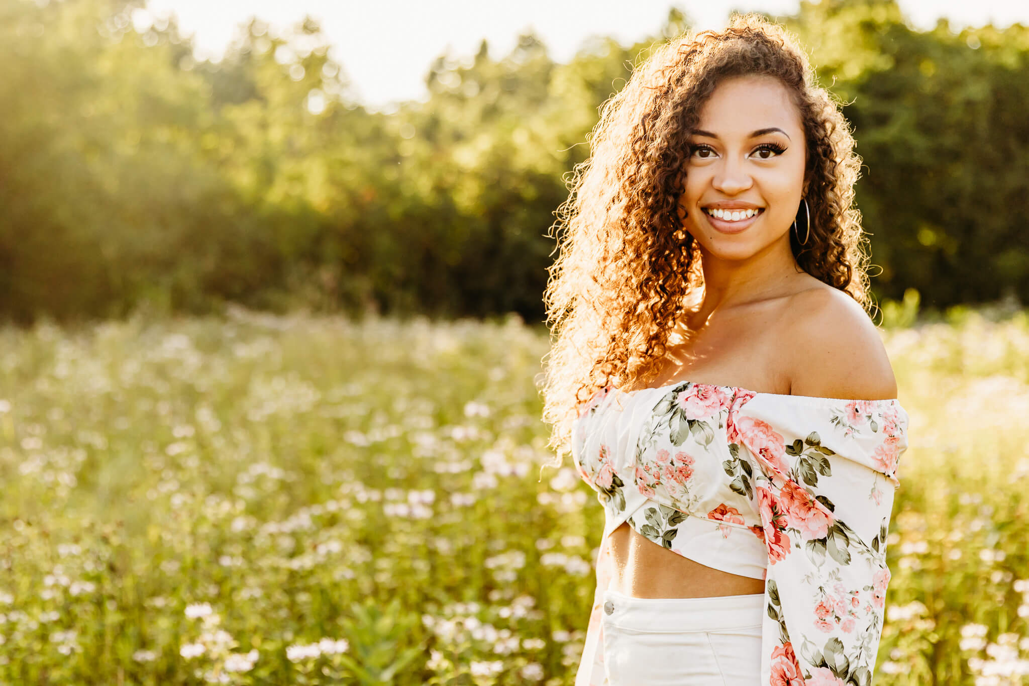 gorgeous high school senior in a floral top and white pants smiling while standing in flowers near Green Bay