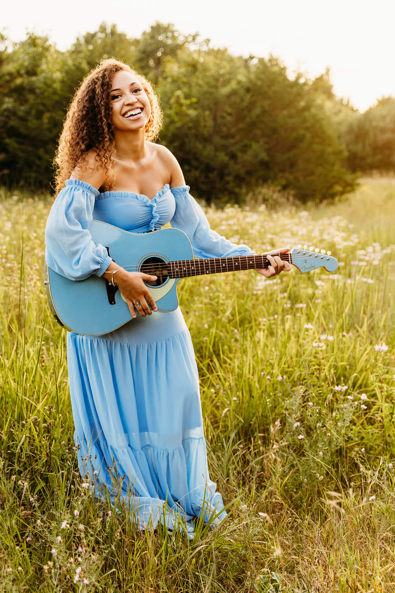 high school senior in a blue dress playing her guitar at sunset in a field