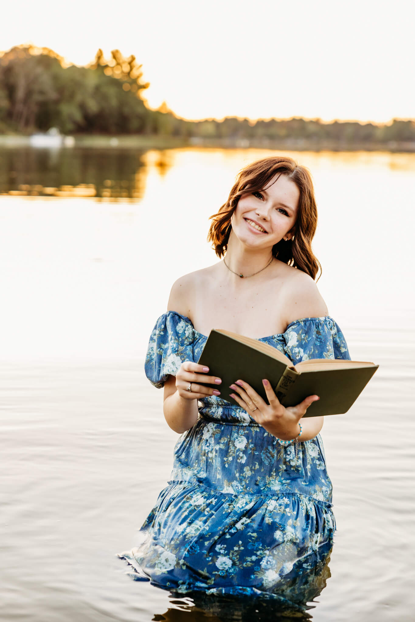 teenage girl in a blue floral dress sitting on a stool in the water while reading a book 