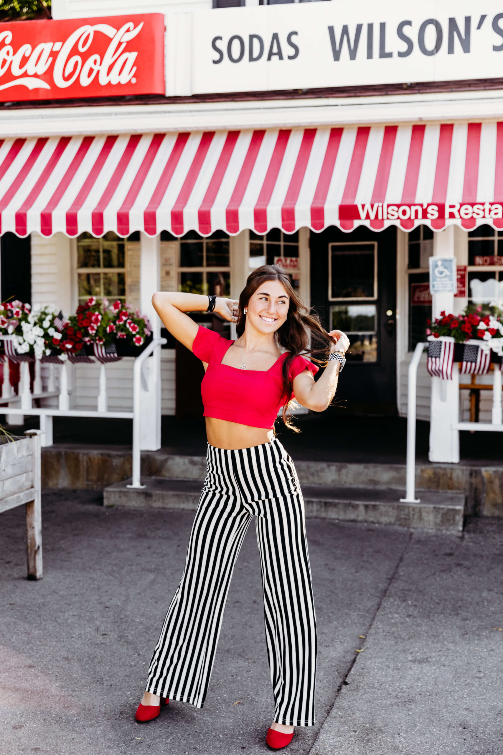 Notre Dame Academy Green Bay senior in a red top with black and white pants playing with her hair in front of Wilsons Restaurant