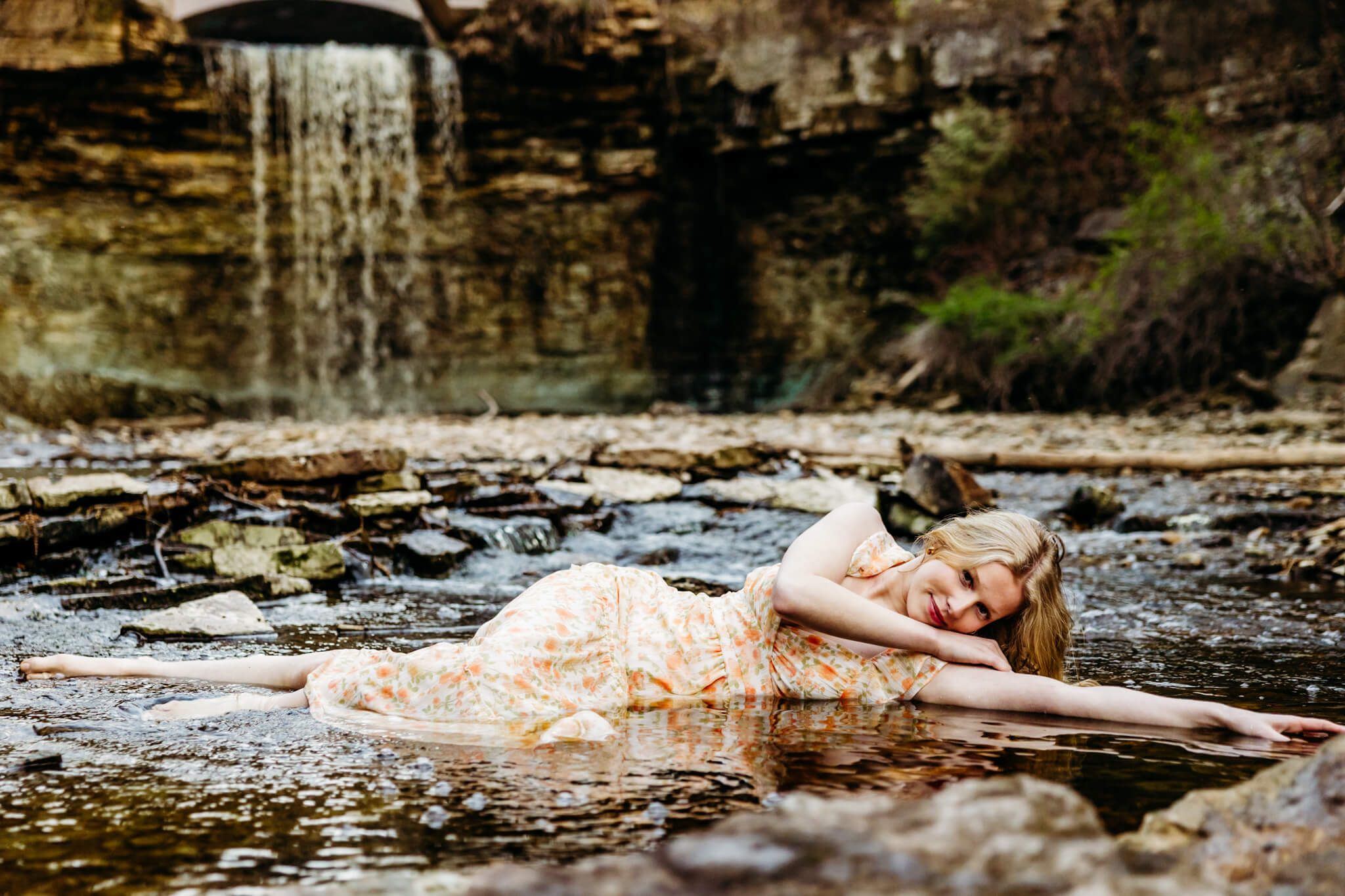gorgeous high school senior girl in a floral dress laying in a creek with a waterfall behind her