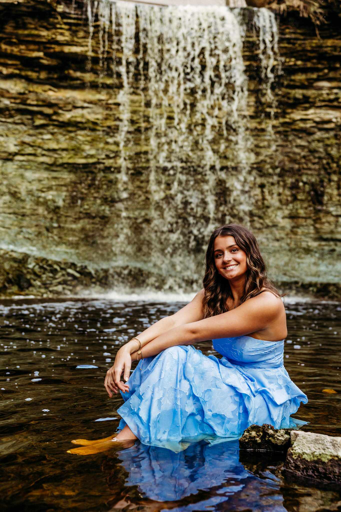 high school senior girl in a blue dress sitting in the water at the base of Wequiock Falls.