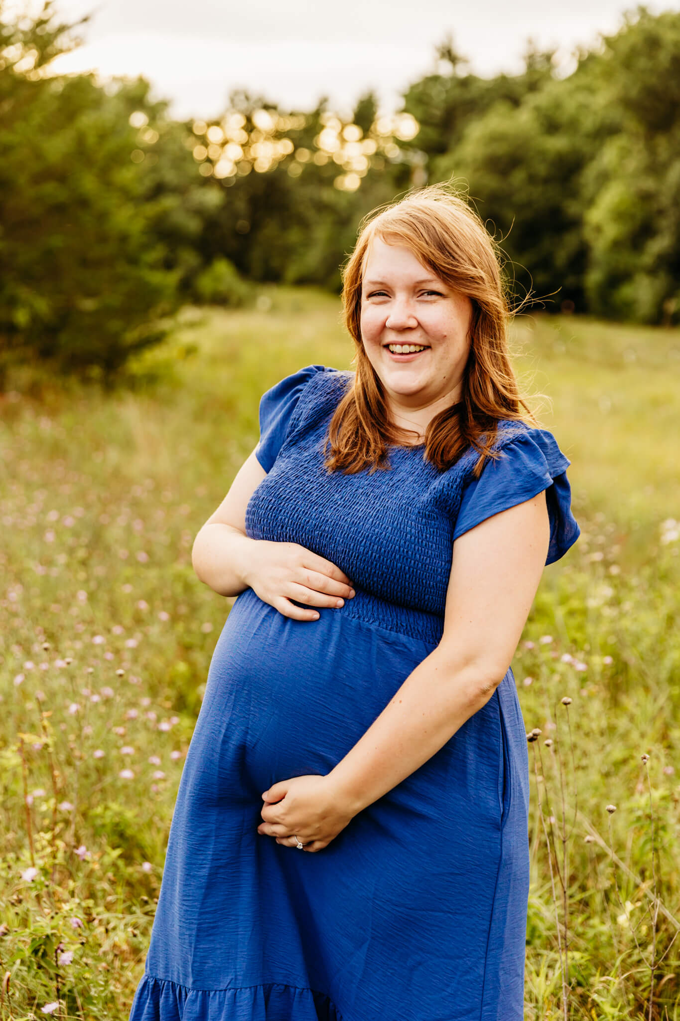 beautiful expecting mom in a blue dress with red hair embracing her bump and laughing during maternity photo session for blog post about green bay massage therapy
