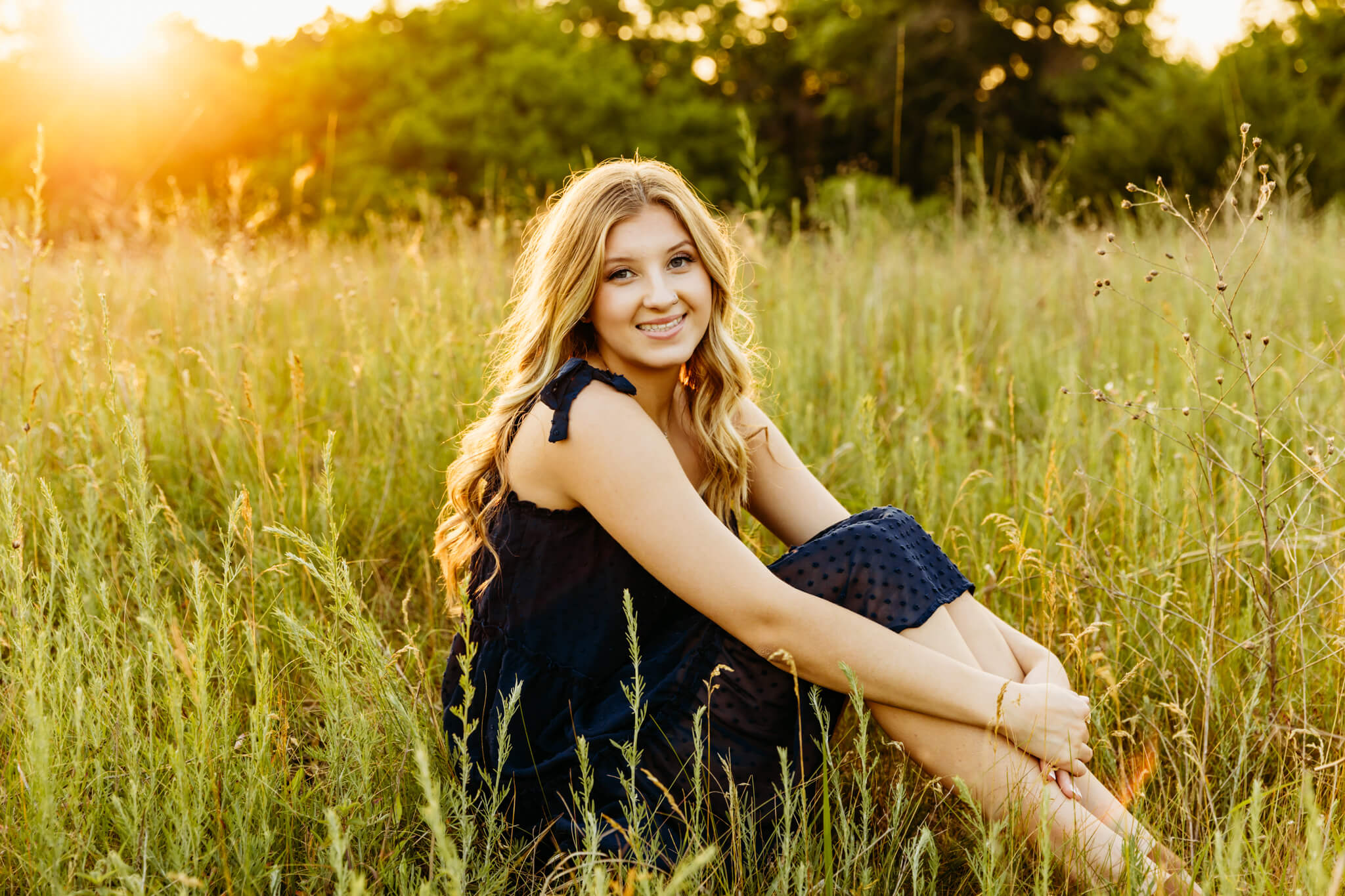beautiful blonde teen girl sitting in tall grass with the sun shining behind her by Ashley Kalbus