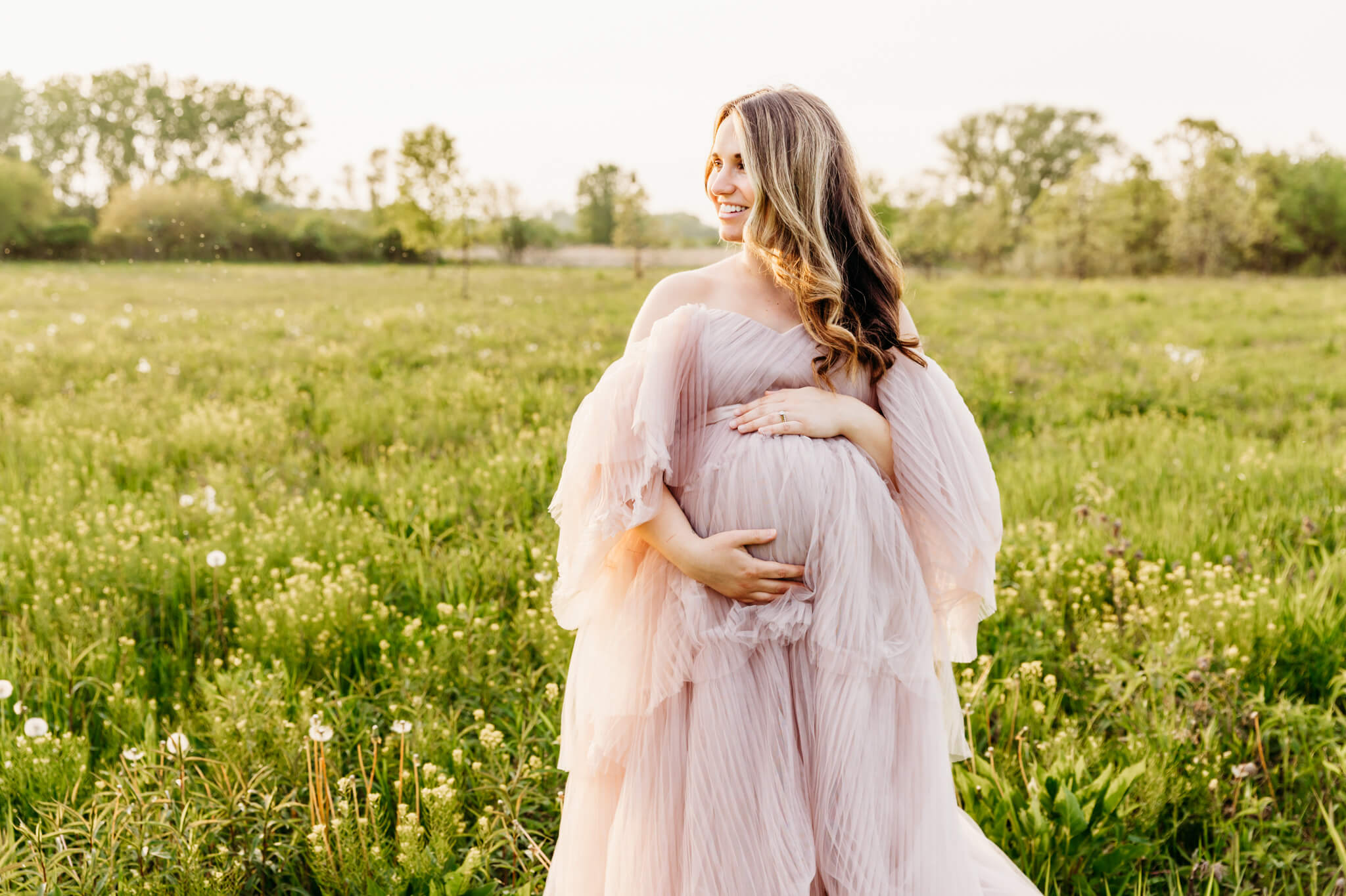 stunning expecting mother in an elaborate blush gown, holds her baby bump and looks into the field