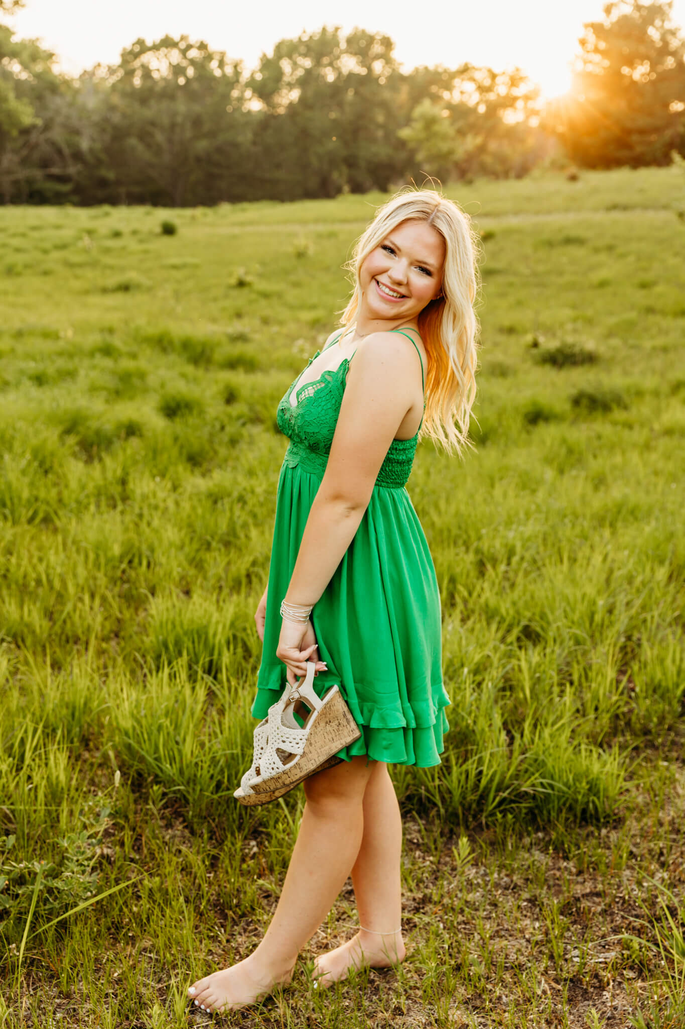 high school teen in a green dress holding her sandals in a field at sunset near Appleton