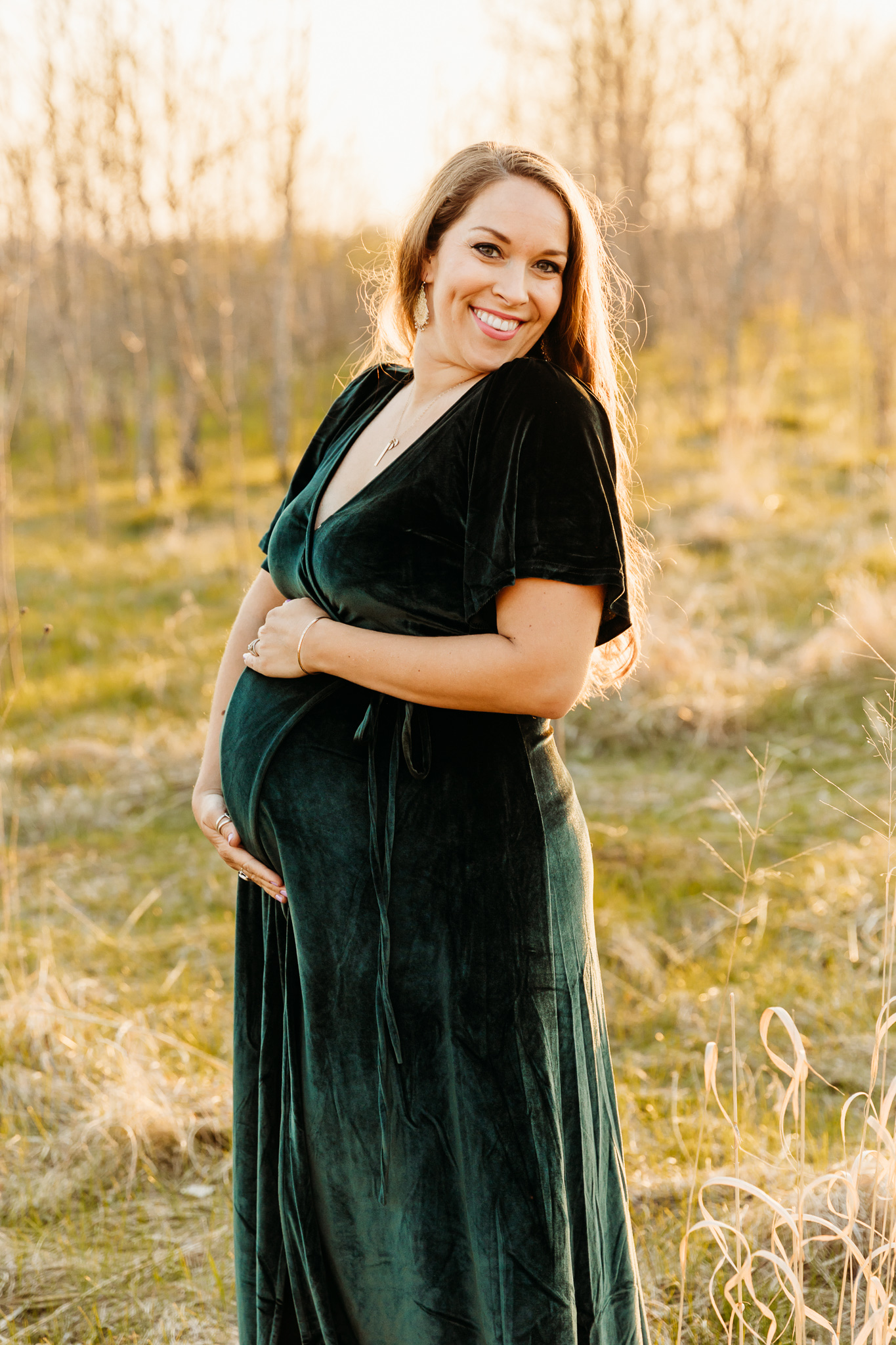 A mom to be holds her bump in a green velvet maternity gown while standing a field at sunset Alba Birth Center