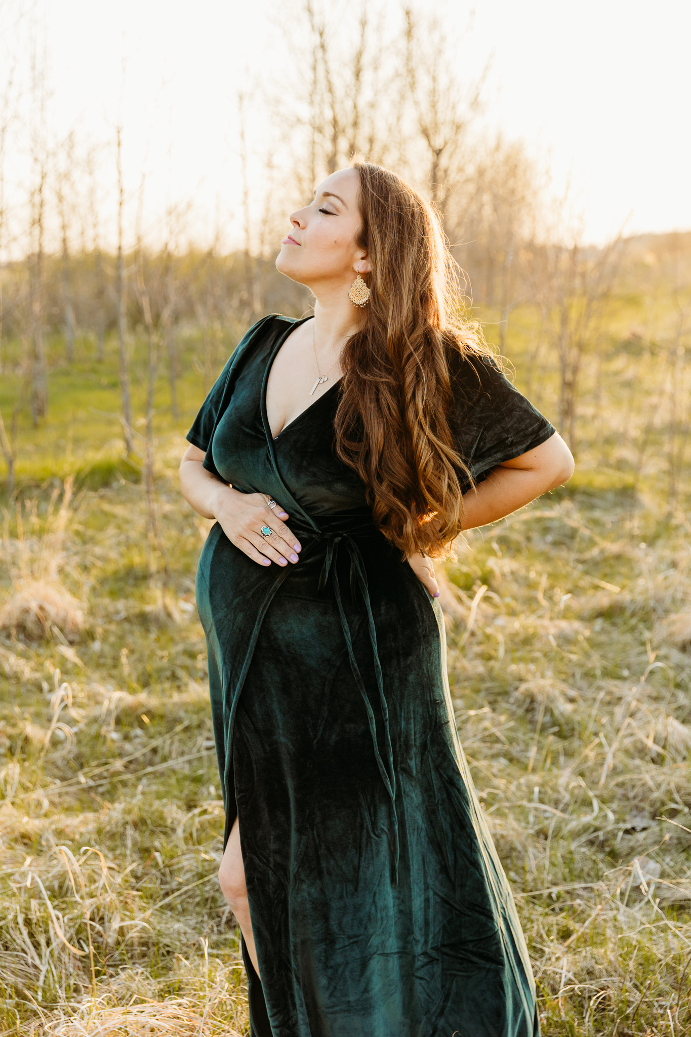 A mother to be stands in a velvet maternity gown in a field of tall grass while soaking in the sun Alba Birth Center