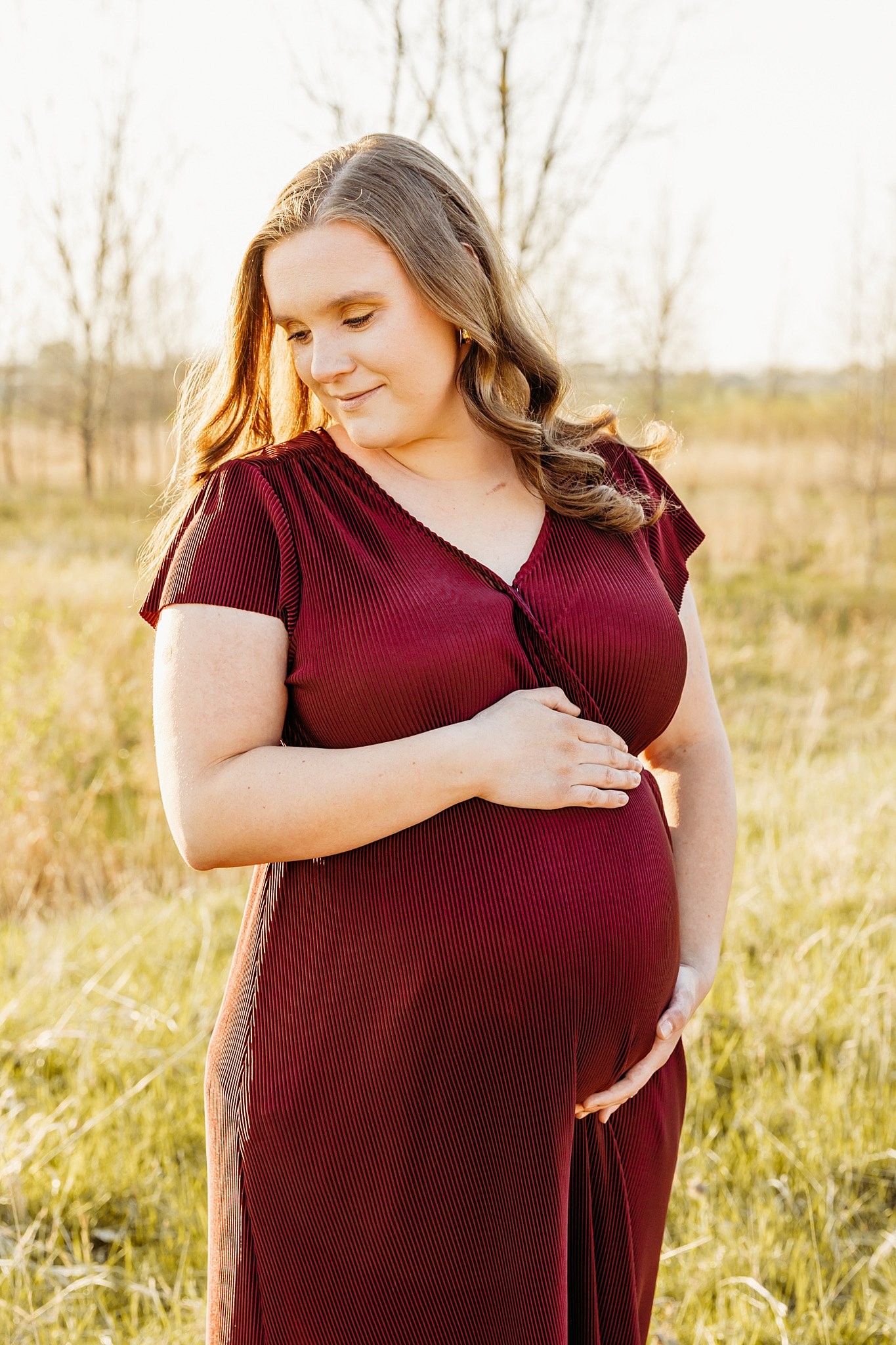 A mother to be in a red maternity gown looks down her shoulder in a field while holding her bump Aurora birthing center