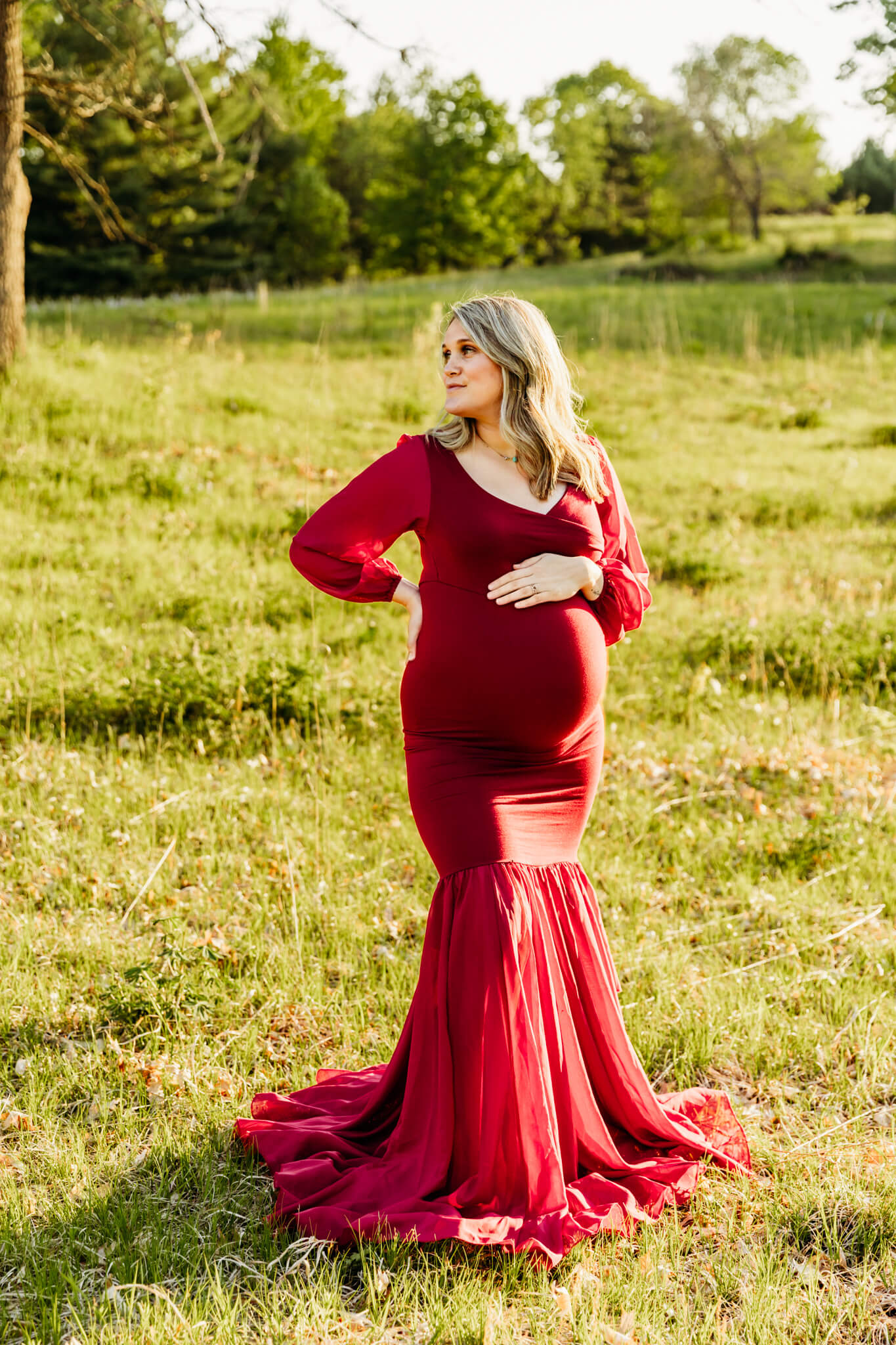 beautiful expecting mom in a fitted red gown resting her hand on her baby bump as she looks over her shoulder in a park