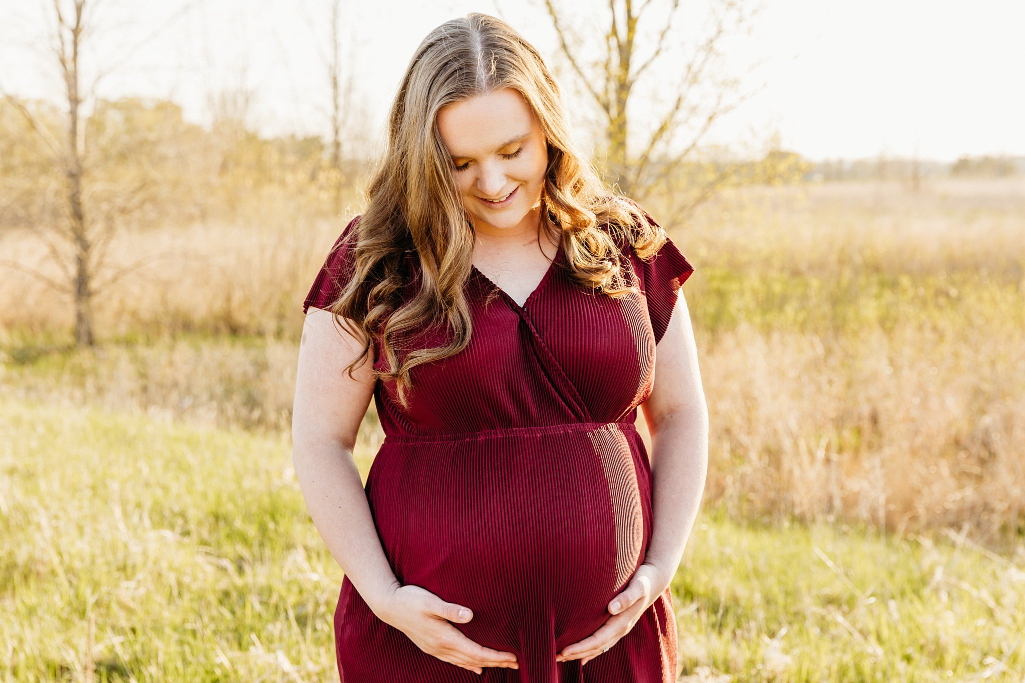 A mother to be in a red maternity dress stands in a field holding her bump Childrens Products LLC