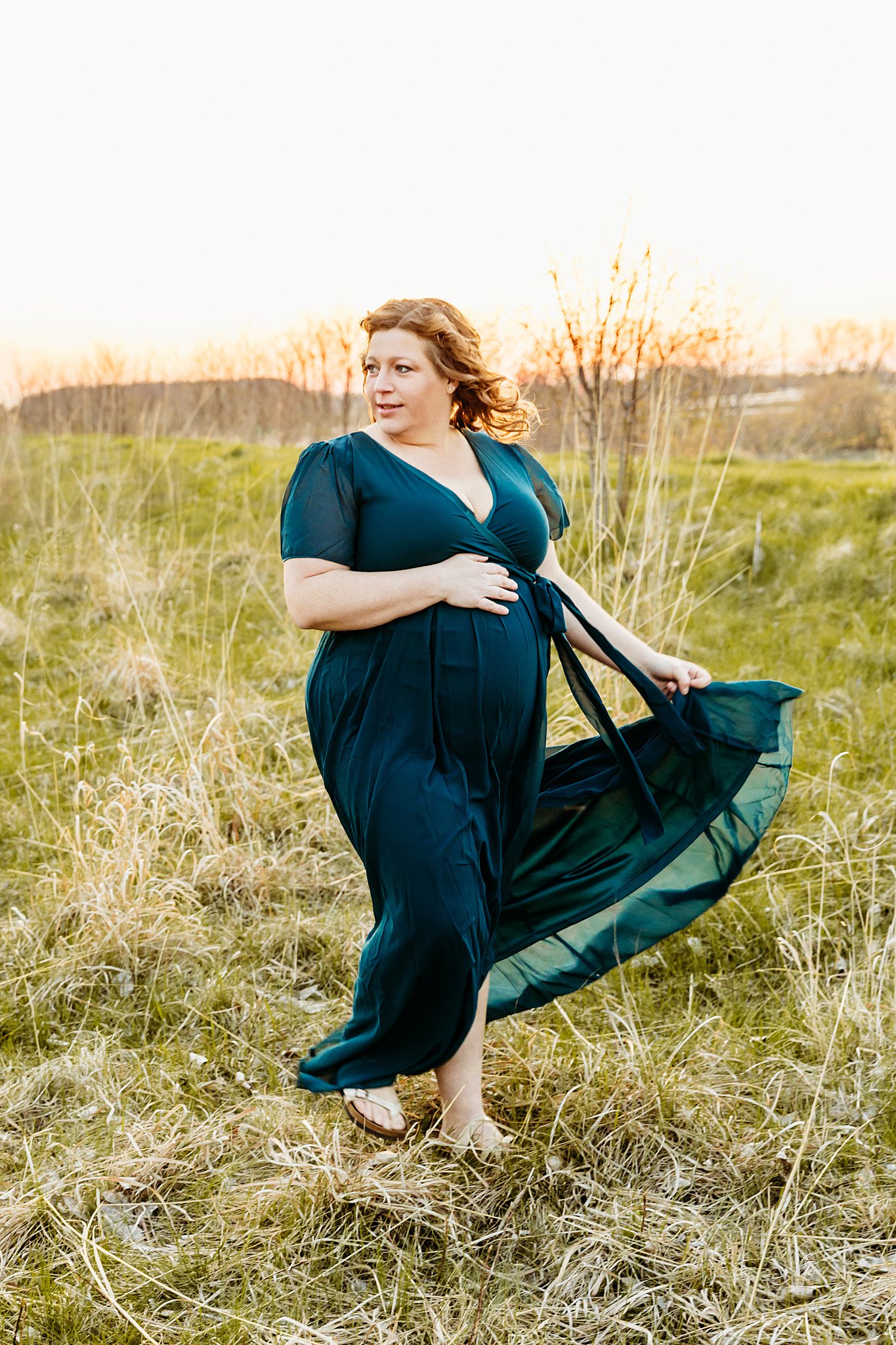 A pregnant woman walks through an open field at sunset while playing with her blue maternity dress Door County Massage