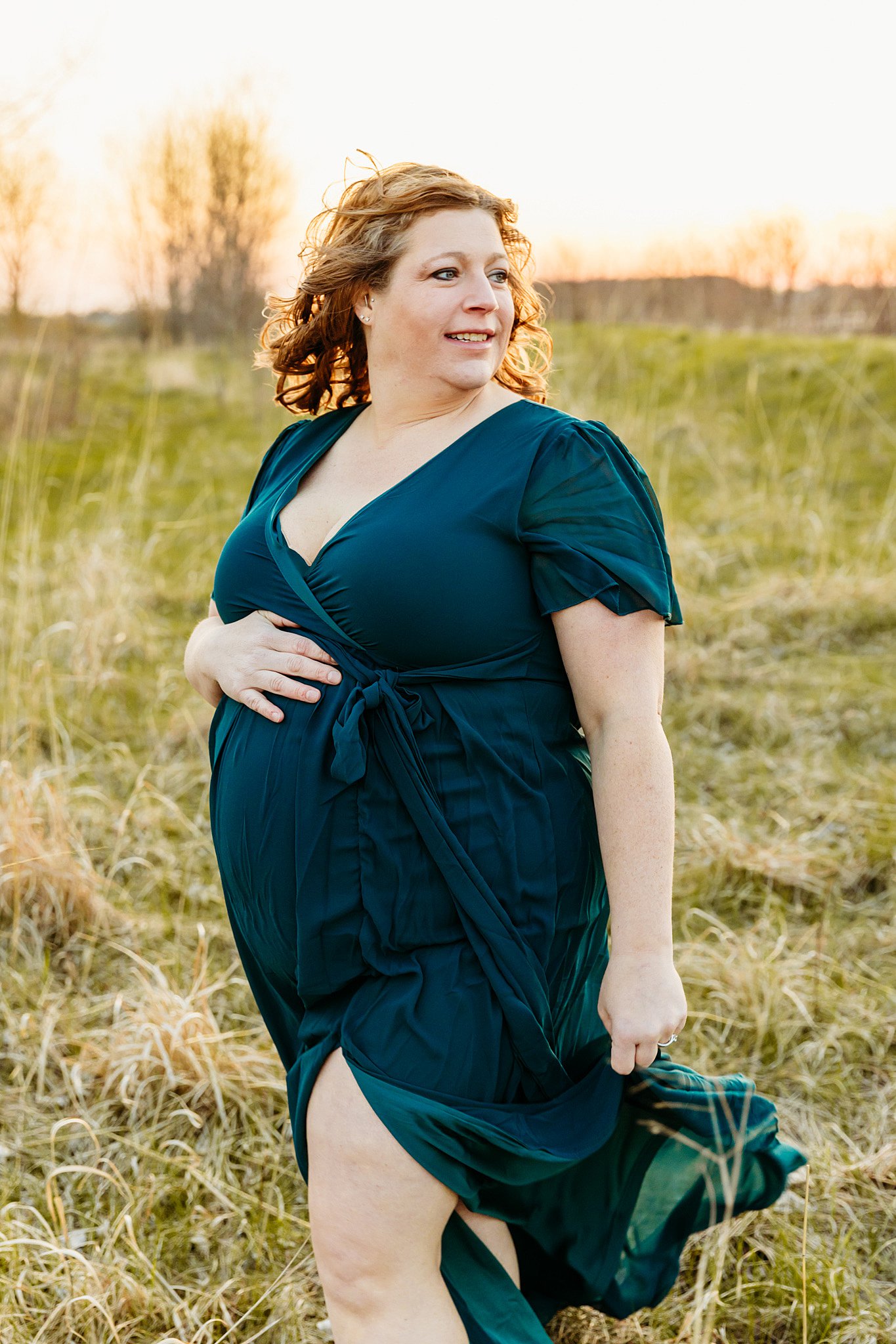 A mother to be in a blue dress that is blowing in the wind stands in an open field at sunset Door County Massage